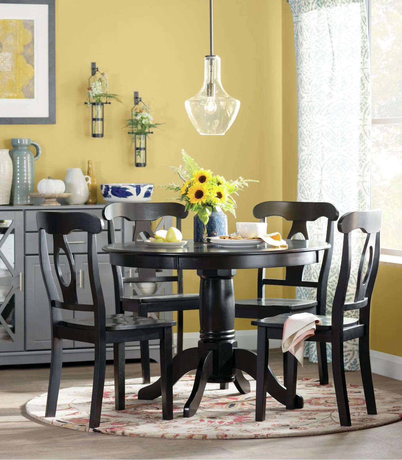 A black round wooden table with four matching chairs sits on a patterned round rug in a dining area with a yellow wall.