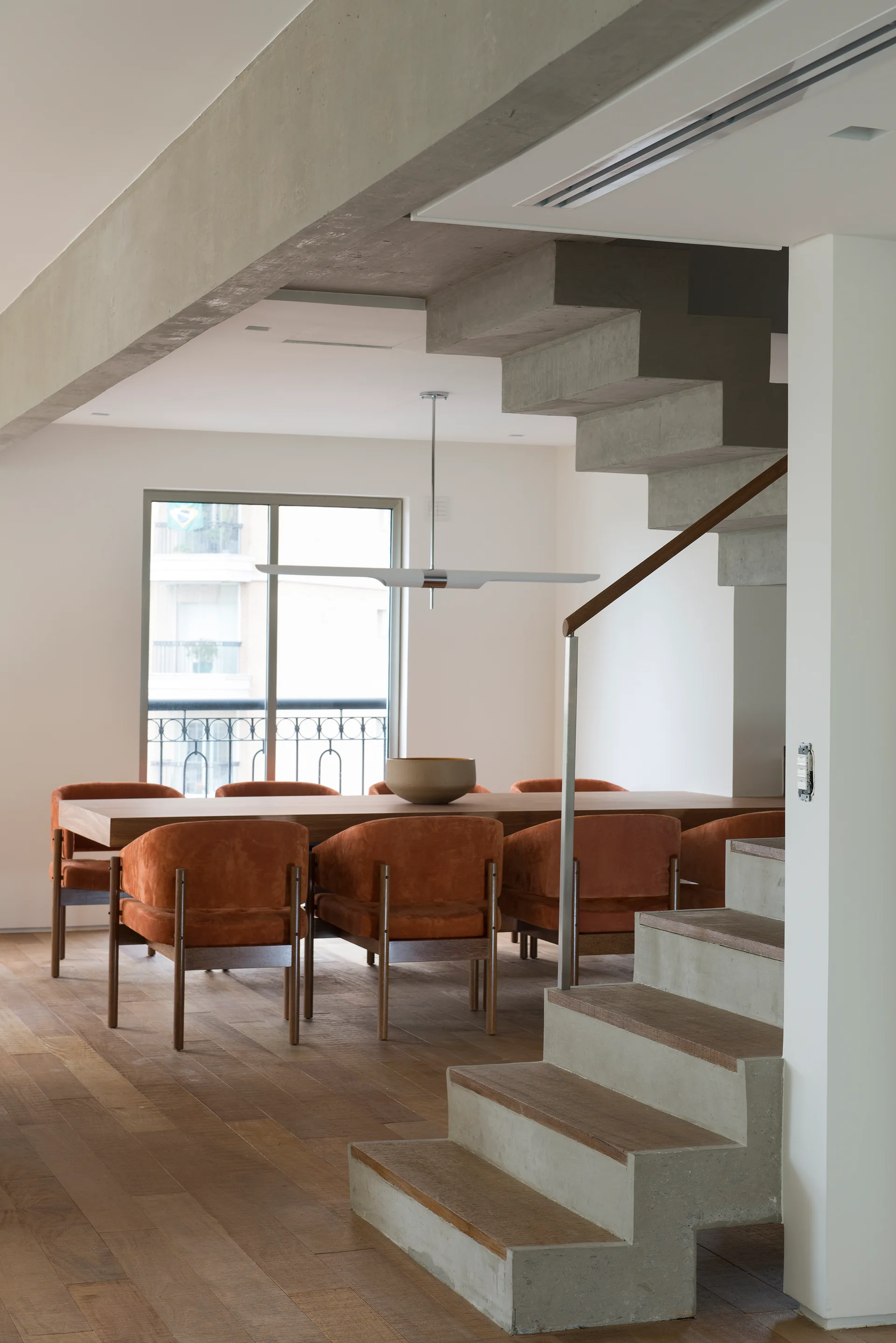 A minimalist dining area featuring a wooden table with burnt orange chairs next to an architectural exposed concrete staircase.