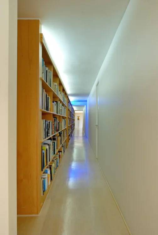 A long, narrow residential hallway features a floor-to-ceiling wooden bookcase packed with books and a glowing cove light illuminating the white ceiling.