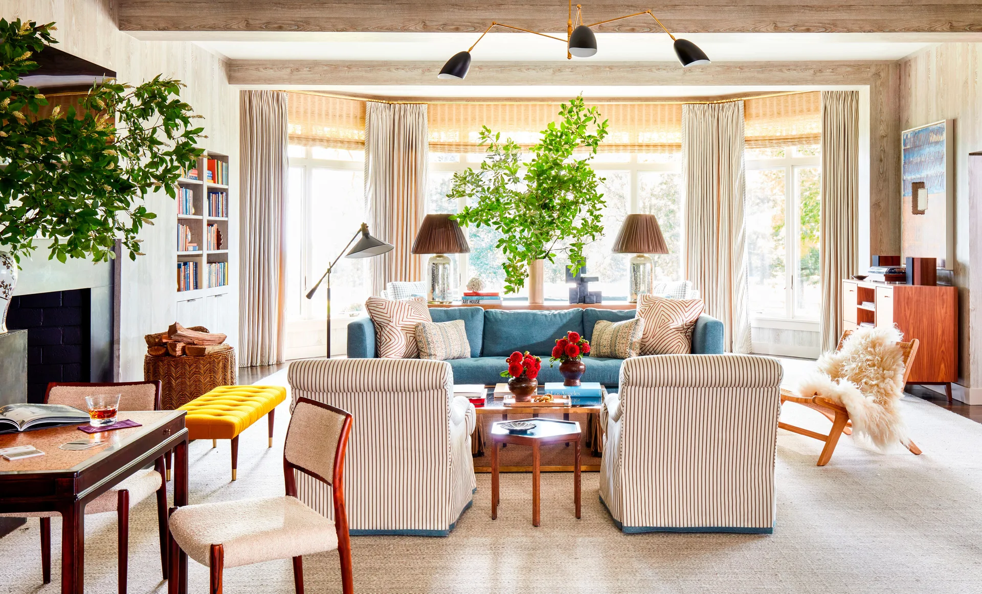 A bright living room features a blue sofa, a pair of striped armchairs, a yellow bench, and wood-beamed ceilings.