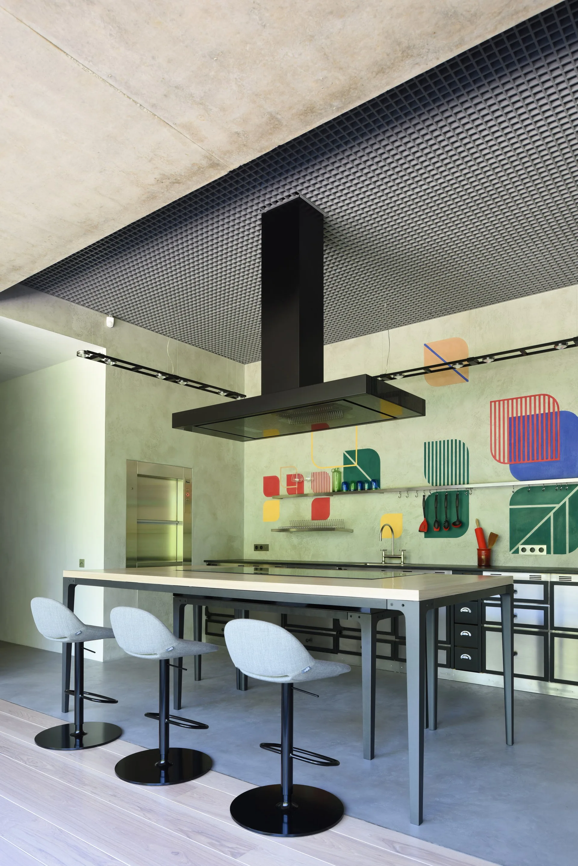 A modern kitchen featuring a central wooden island with grey stools under a large black range hood, with colorful geometric art on a light green wall.