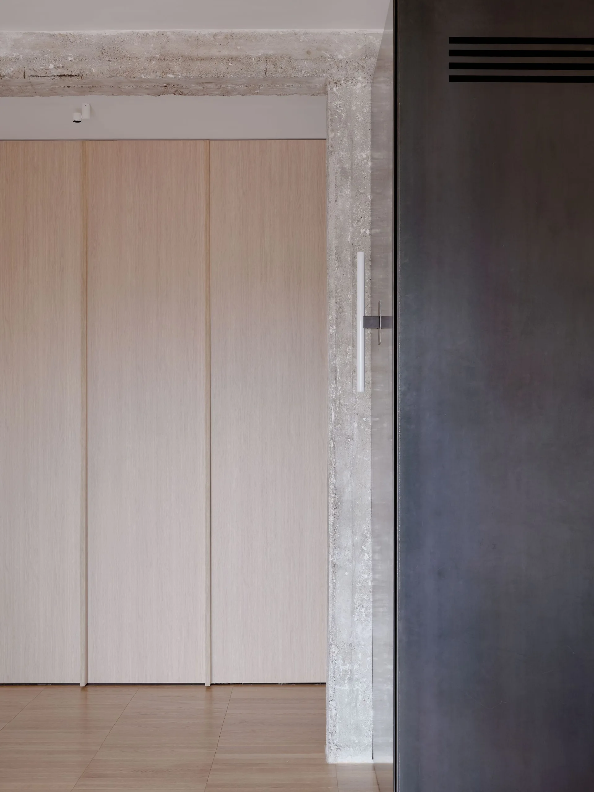 Full-height light wood storage panels stand adjacent to a rough, exposed concrete column featuring a vertical wall sconce and a dark metal panel.