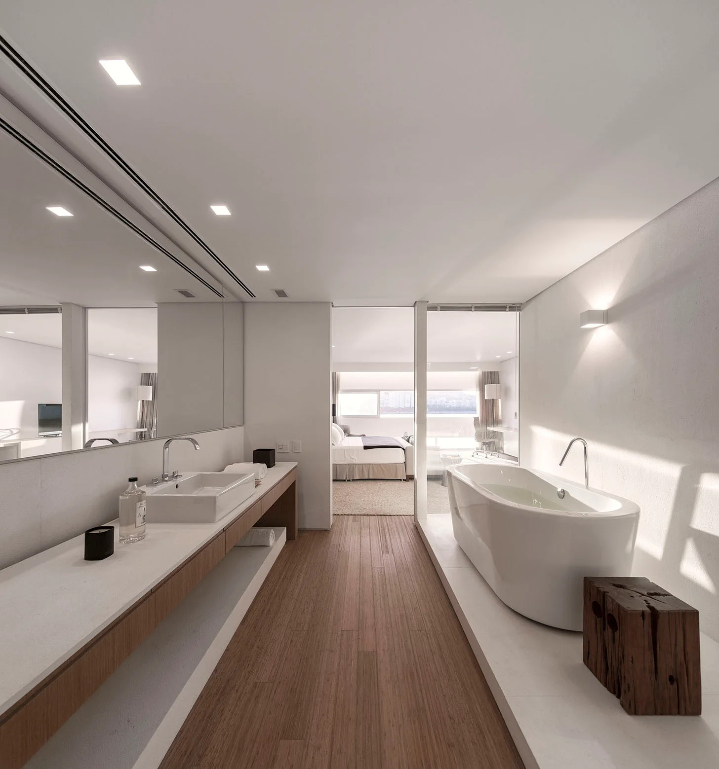 A minimalist bathroom featuring a long floating vanity with a wide mirror, a white freestanding bathtub, and a wood plank floor.