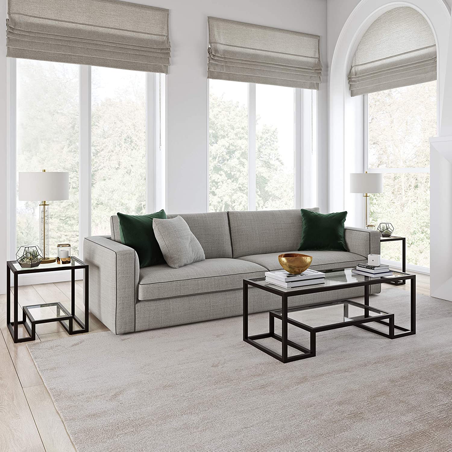 A gray sofa with dark green throw pillows sits in a bright living room with geometric black metal and glass tables and large windows.