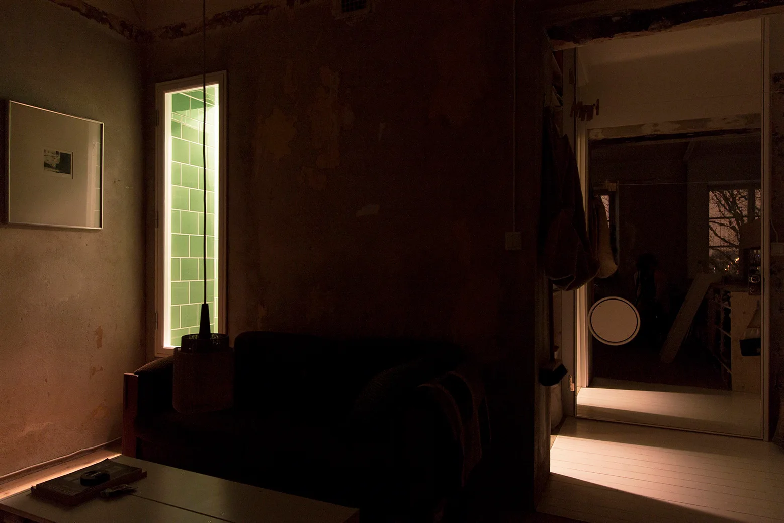 A dimly lit living room with a dark sofa against a rough plaster wall, featuring a narrow glowing window lined with green tiles.