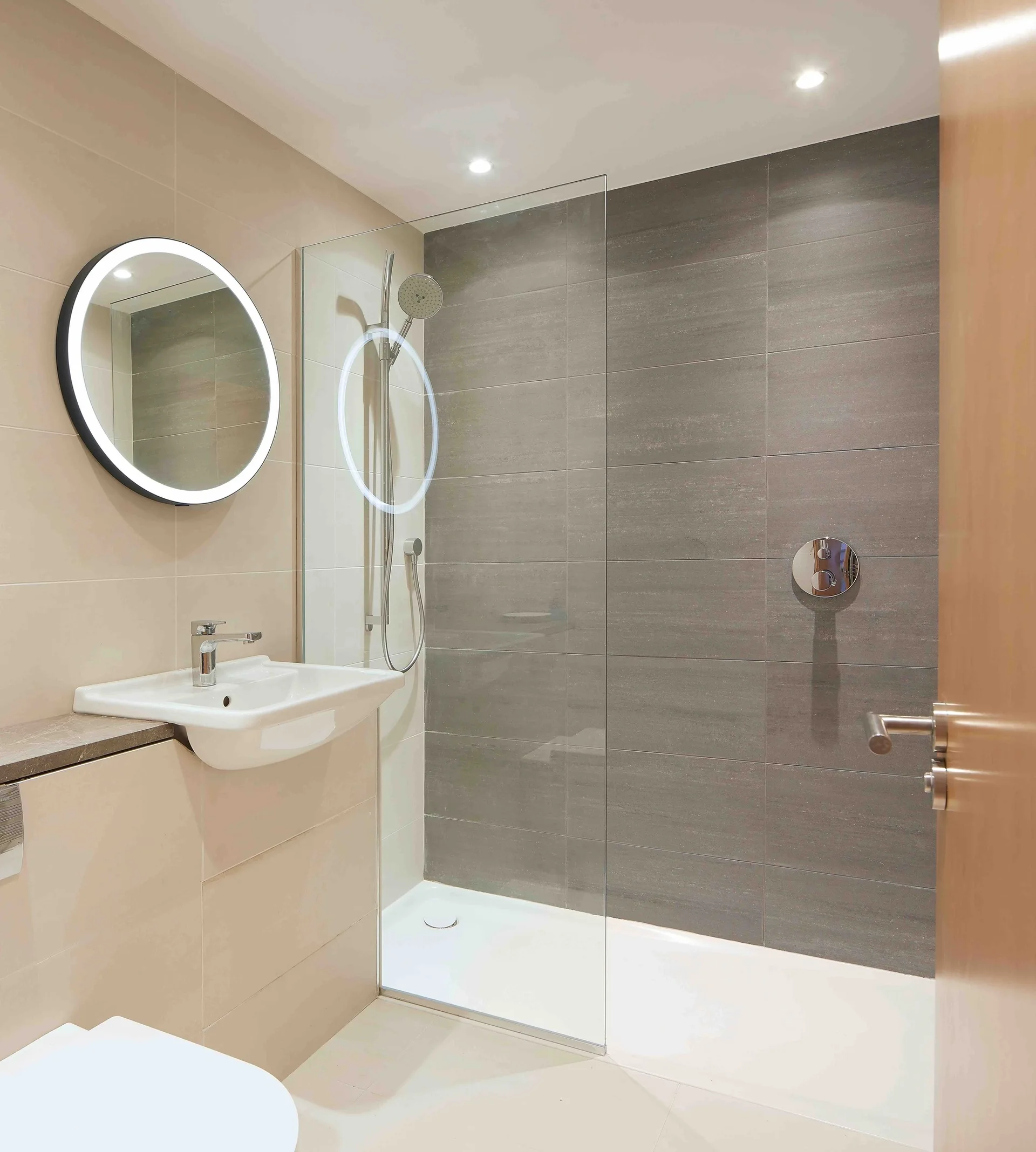 A modern bathroom with a glass-enclosed shower featuring dark grey wall tiles, a beige vanity unit, and a round backlit LED mirror.