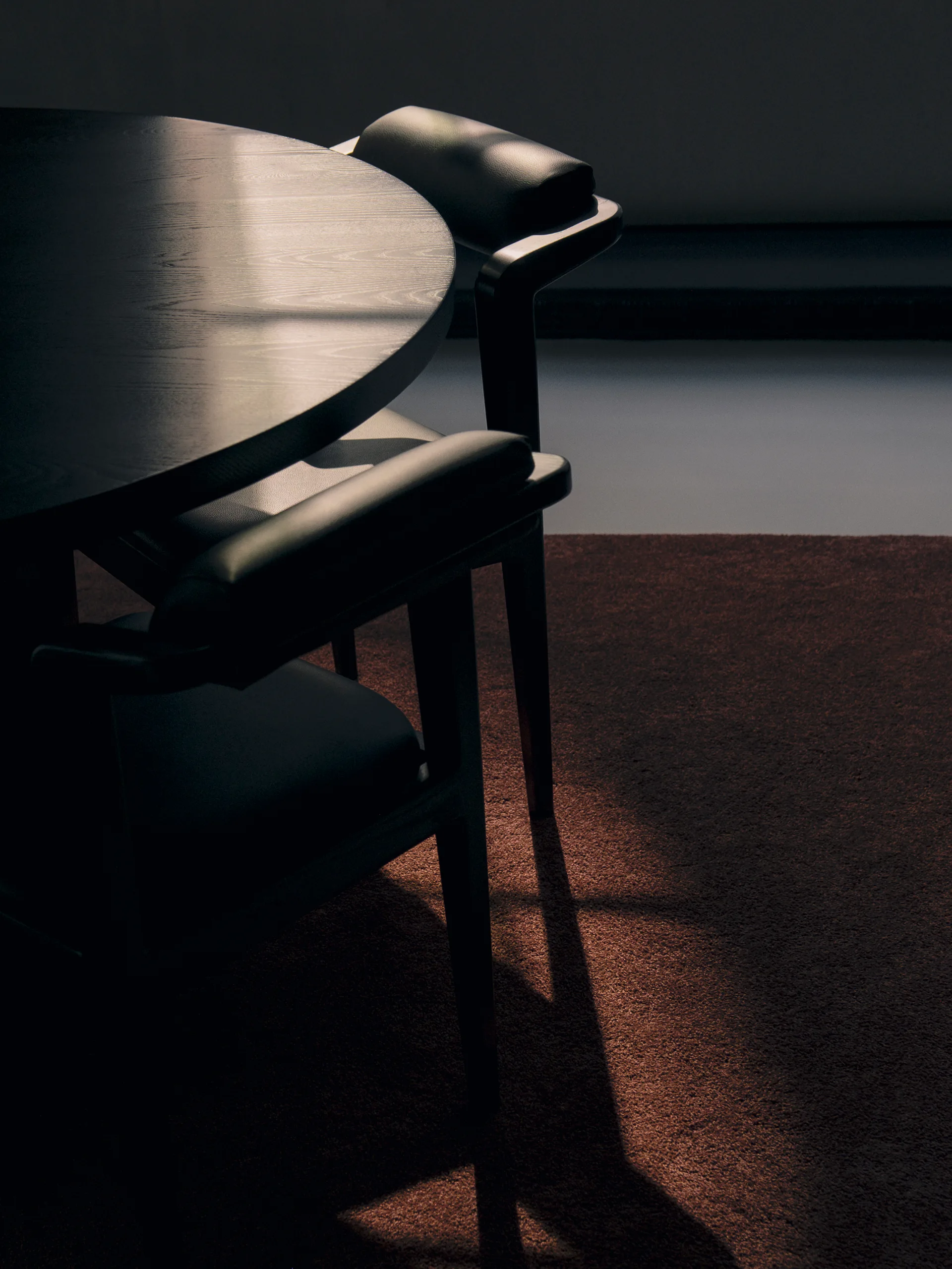 A dark circular wood dining table and two black upholstered chairs sit on a plush brown carpet.