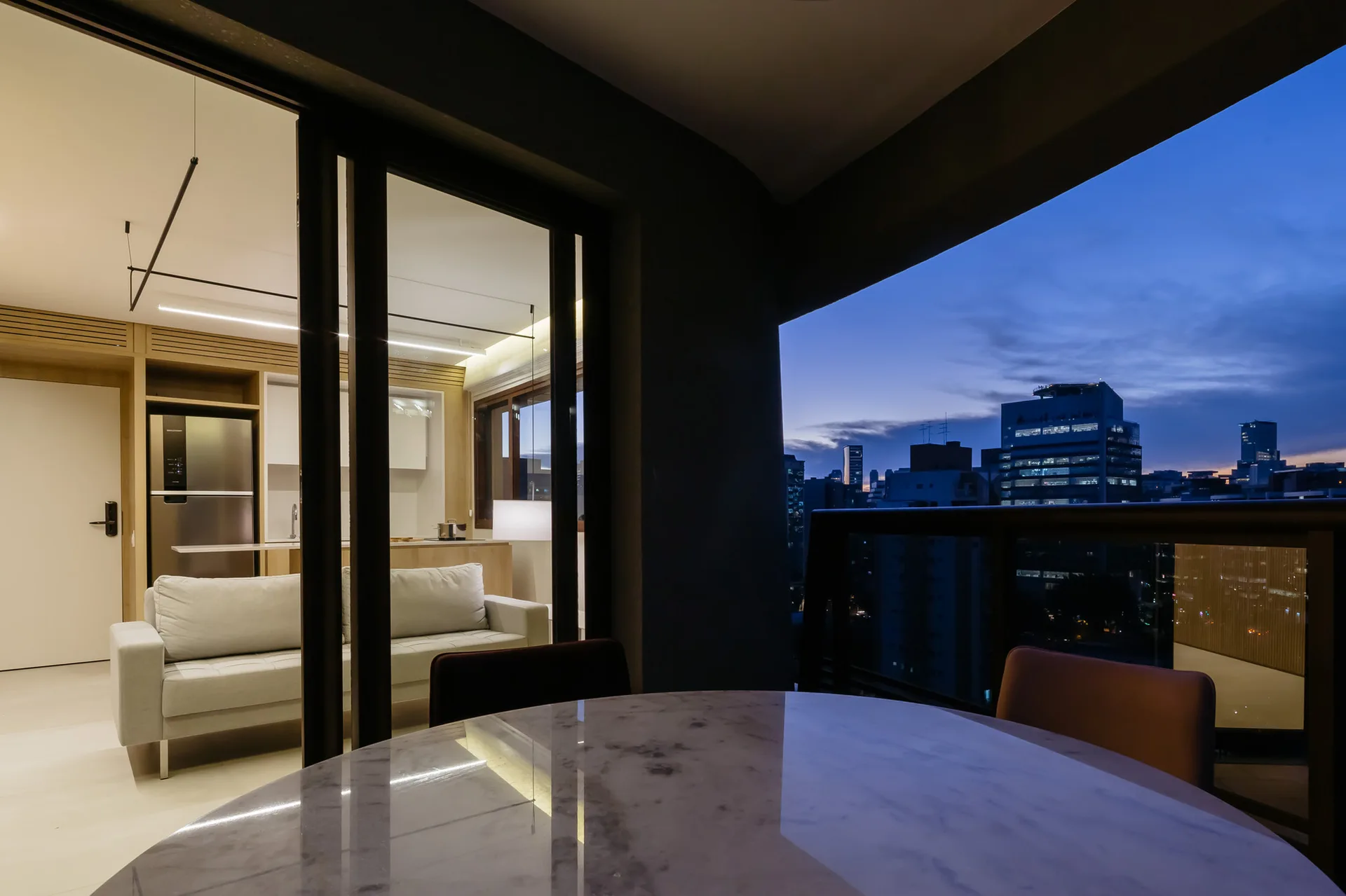 A view from an outdoor balcony with a round marble table looking into a modern, lit apartment living room and kitchen at dusk.