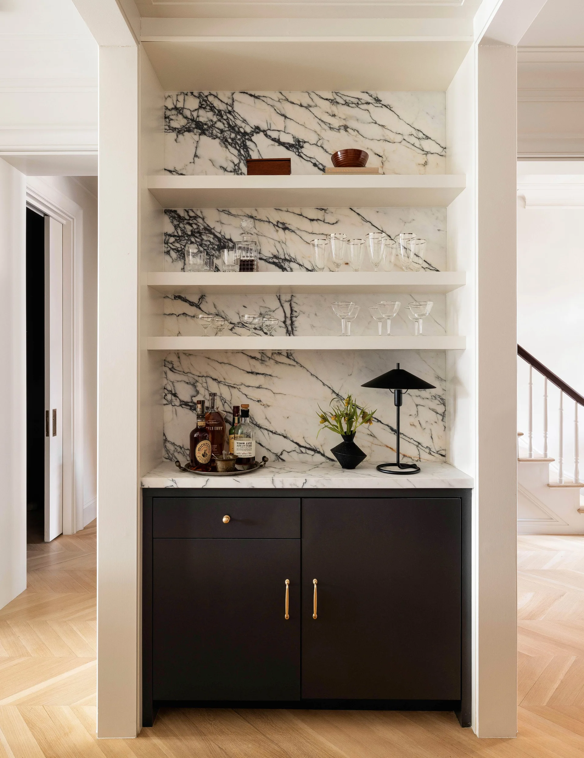 A built-in home bar niche features a black lower cabinet, white veined marble countertops and backsplash, and floating marble shelves with glassware.