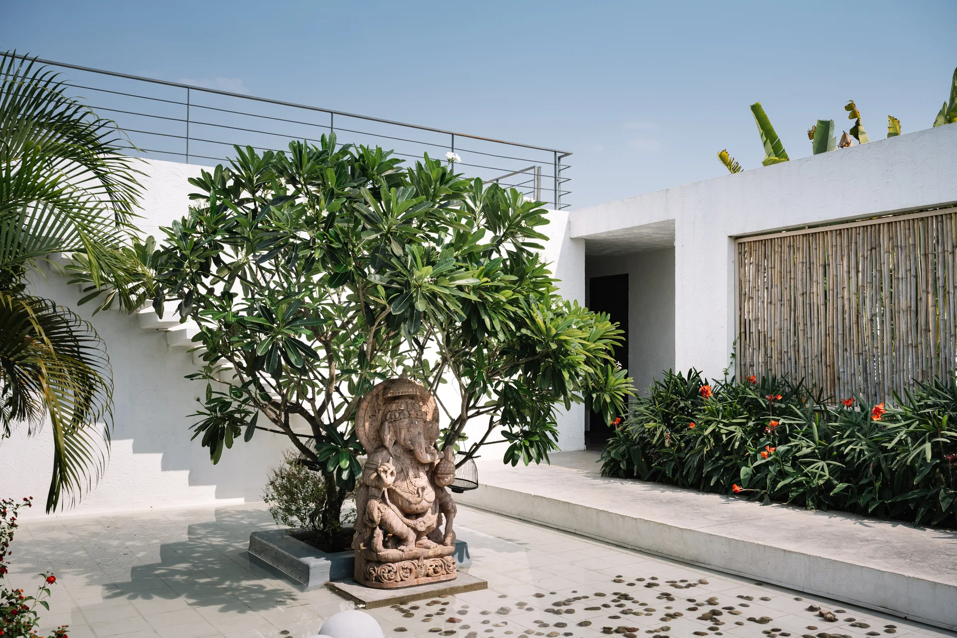 A carved stone statue stands beneath a green tree in a minimalist white-walled courtyard featuring a bamboo privacy screen.