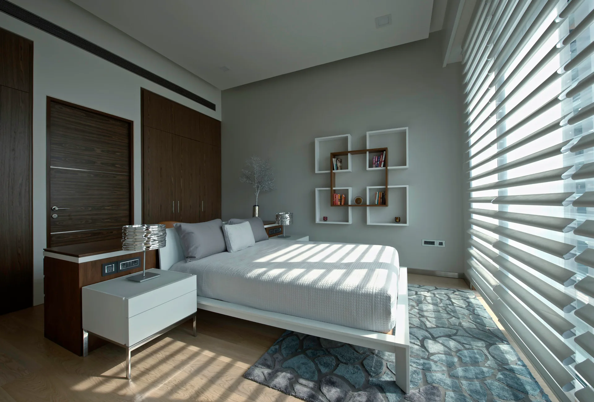 A modern bedroom features a white bed, square wall shelves with books, dark wood wardrobes, and light filtering through horizontal window blinds.