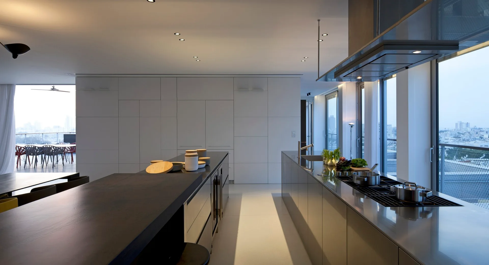 A minimalist kitchen features a long stainless steel island with an integrated cooktop and sink beside a seamless wall of handleless white cabinets.