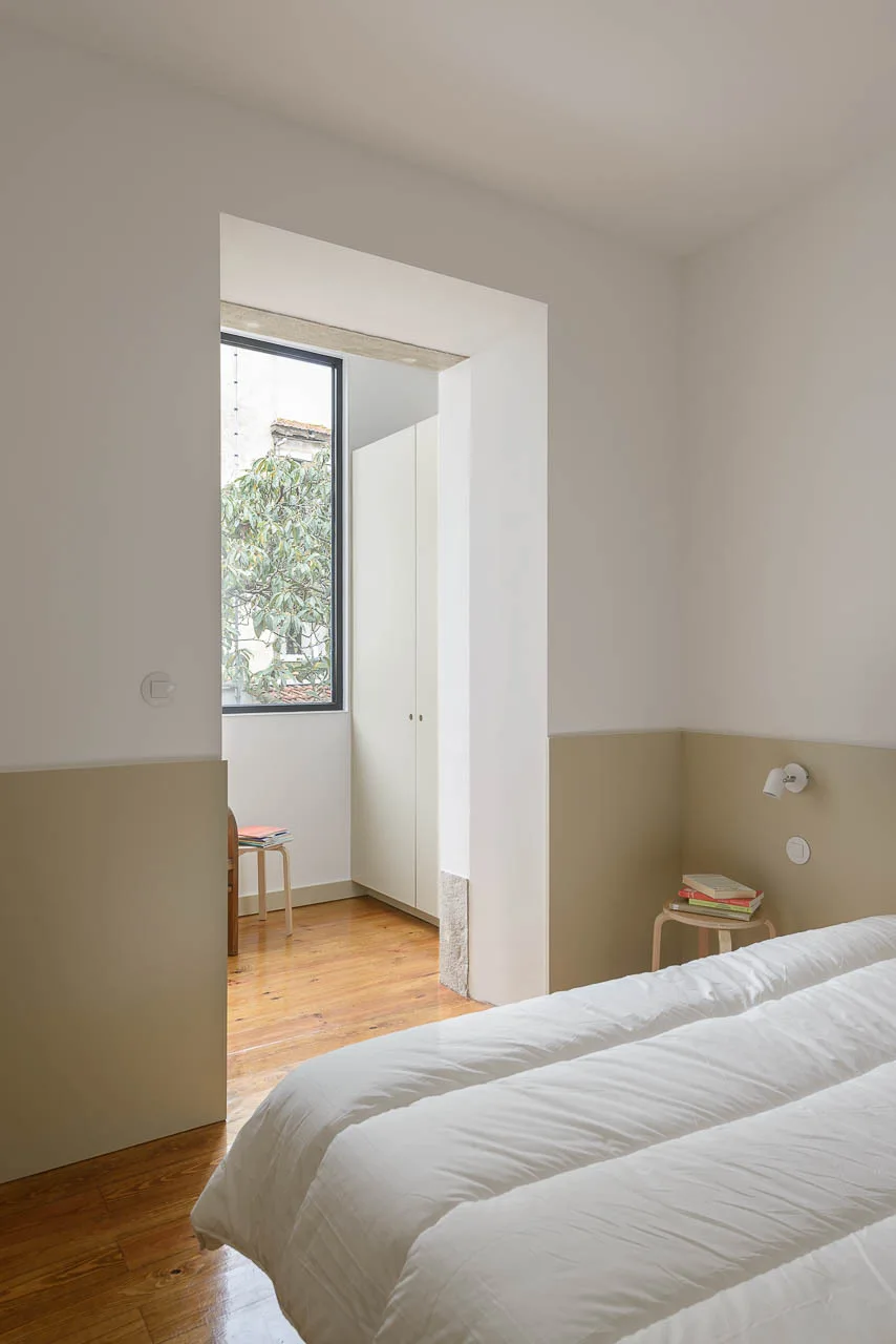 A minimalist bedroom featuring beige color-blocked lower walls, white bedding on a low bed, a wooden stool nightstand, and an opening to a wardrobe area.