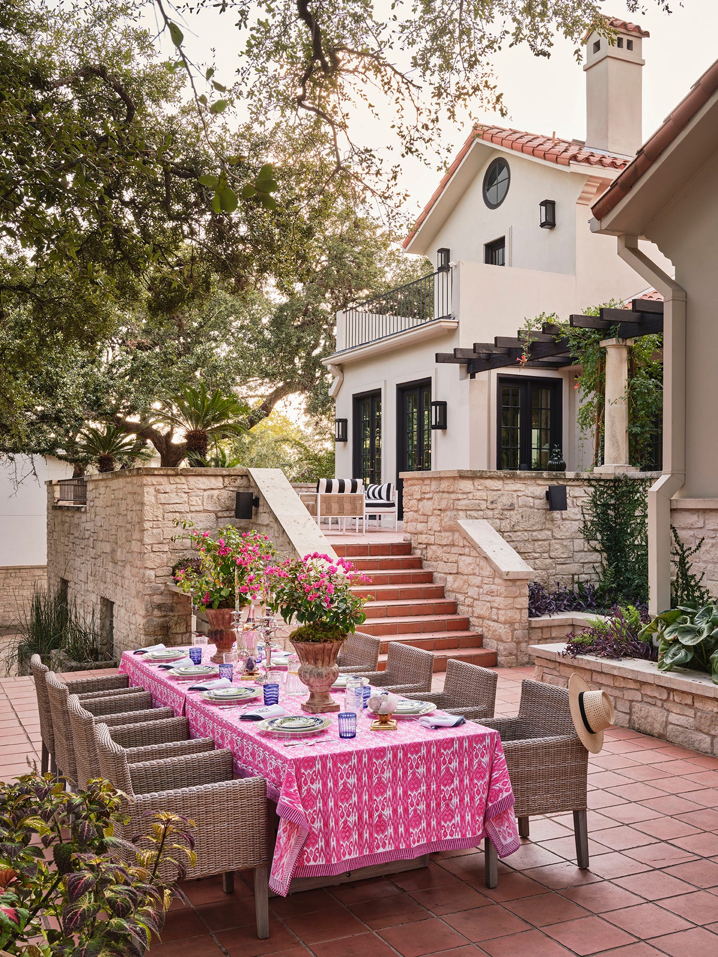 A long outdoor dining table covered in a pink patterned tablecloth is surrounded by woven grey chairs on a terracotta tiled patio next to stone walls.