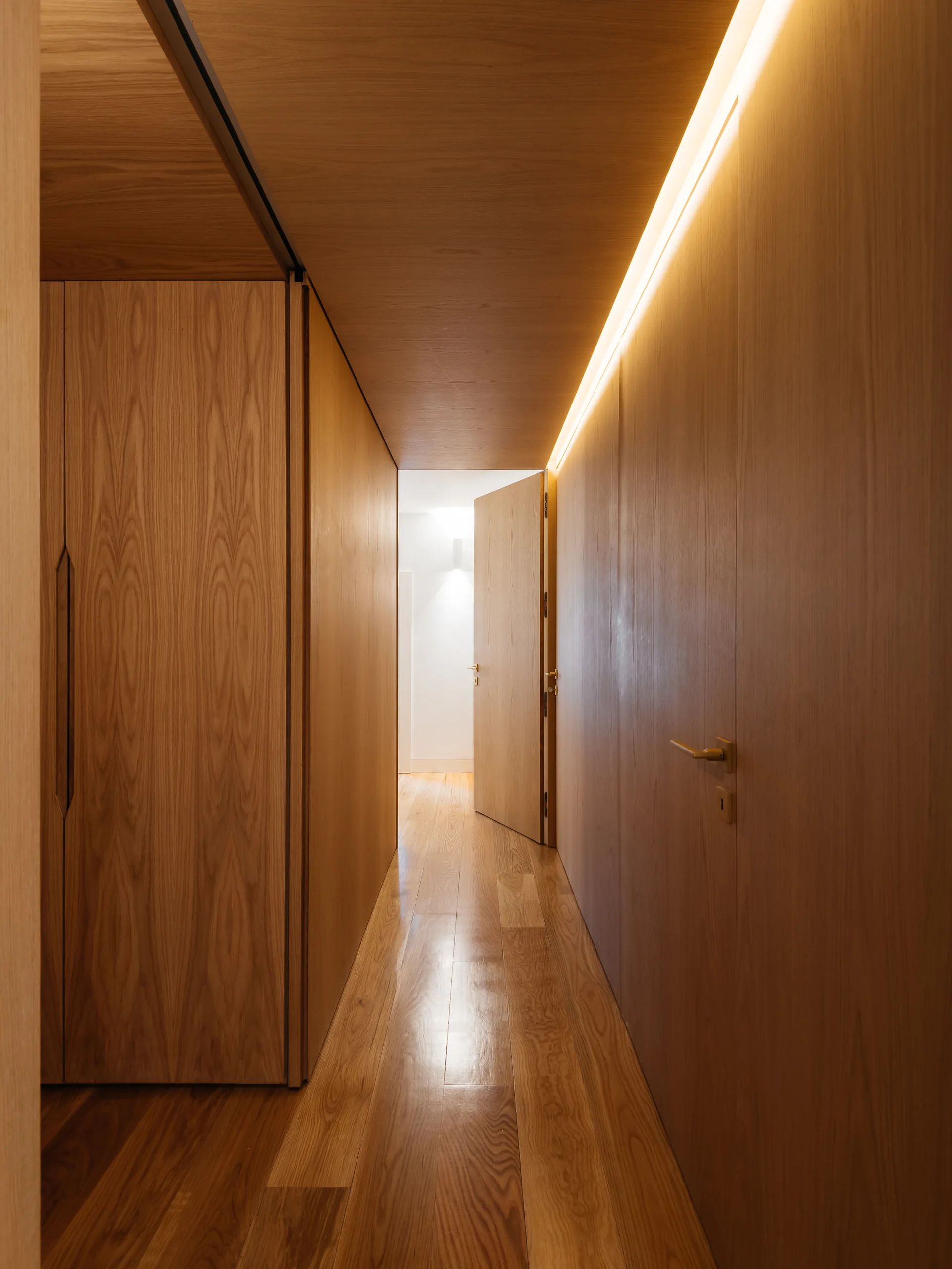 A narrow residential hallway wrapped in warm wood paneling with timber floors, illuminated by a strip of cove lighting along the ceiling.