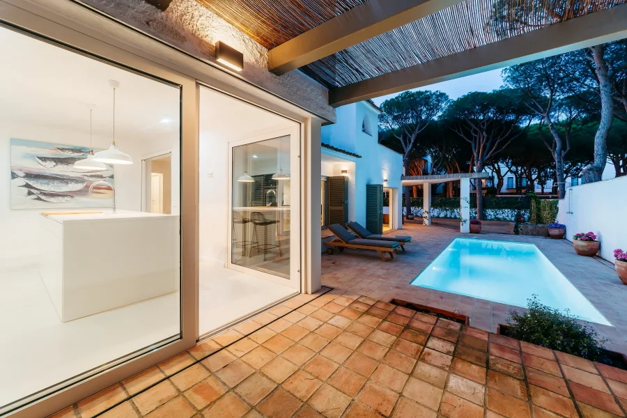 A terracotta tiled patio features a swimming pool alongside sliding glass doors that open to a modern white kitchen.
