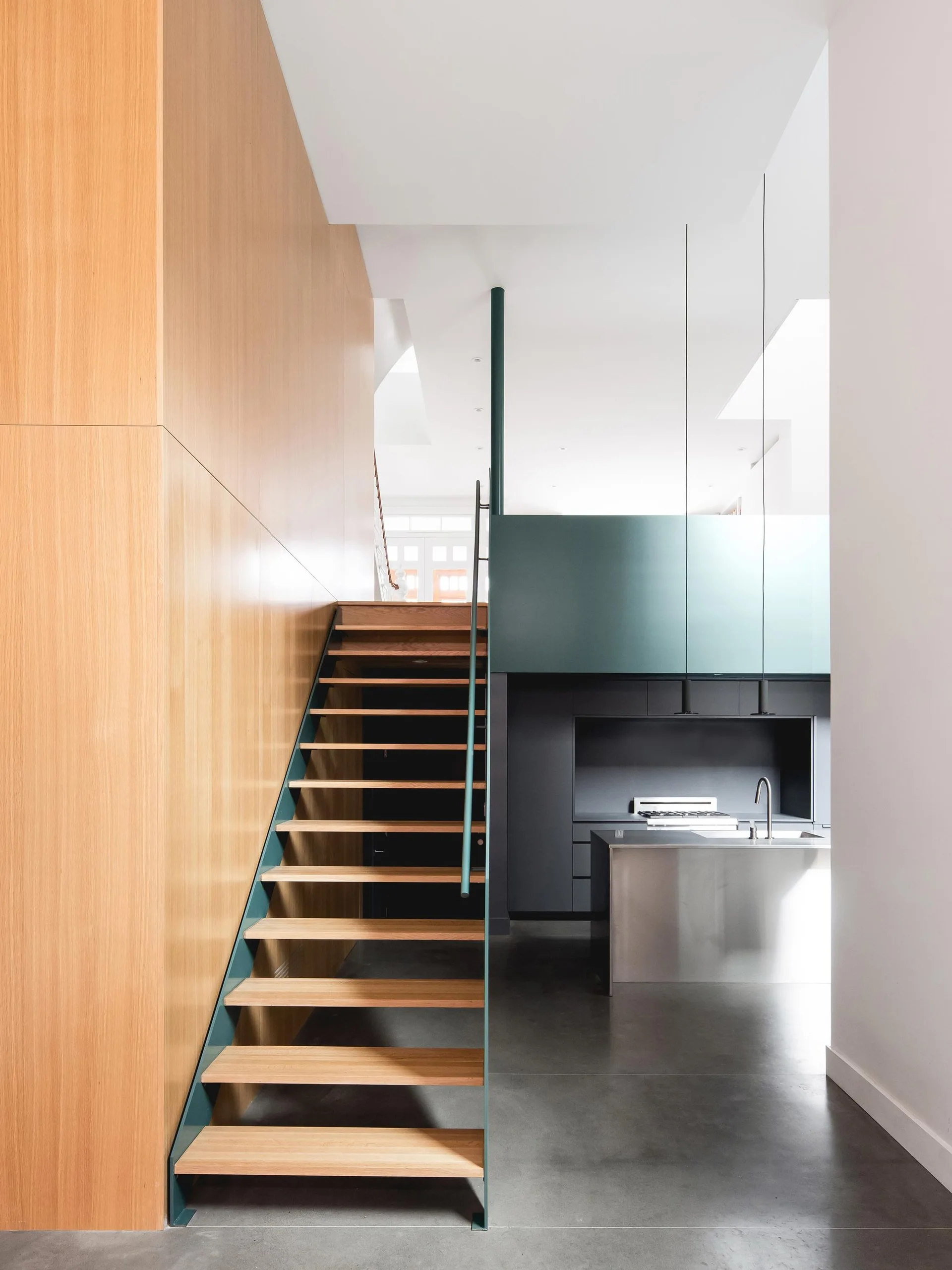 An open-riser wood staircase with teal metal trim sits beside a wood-paneled wall, leading toward a kitchen with a stainless steel island.
