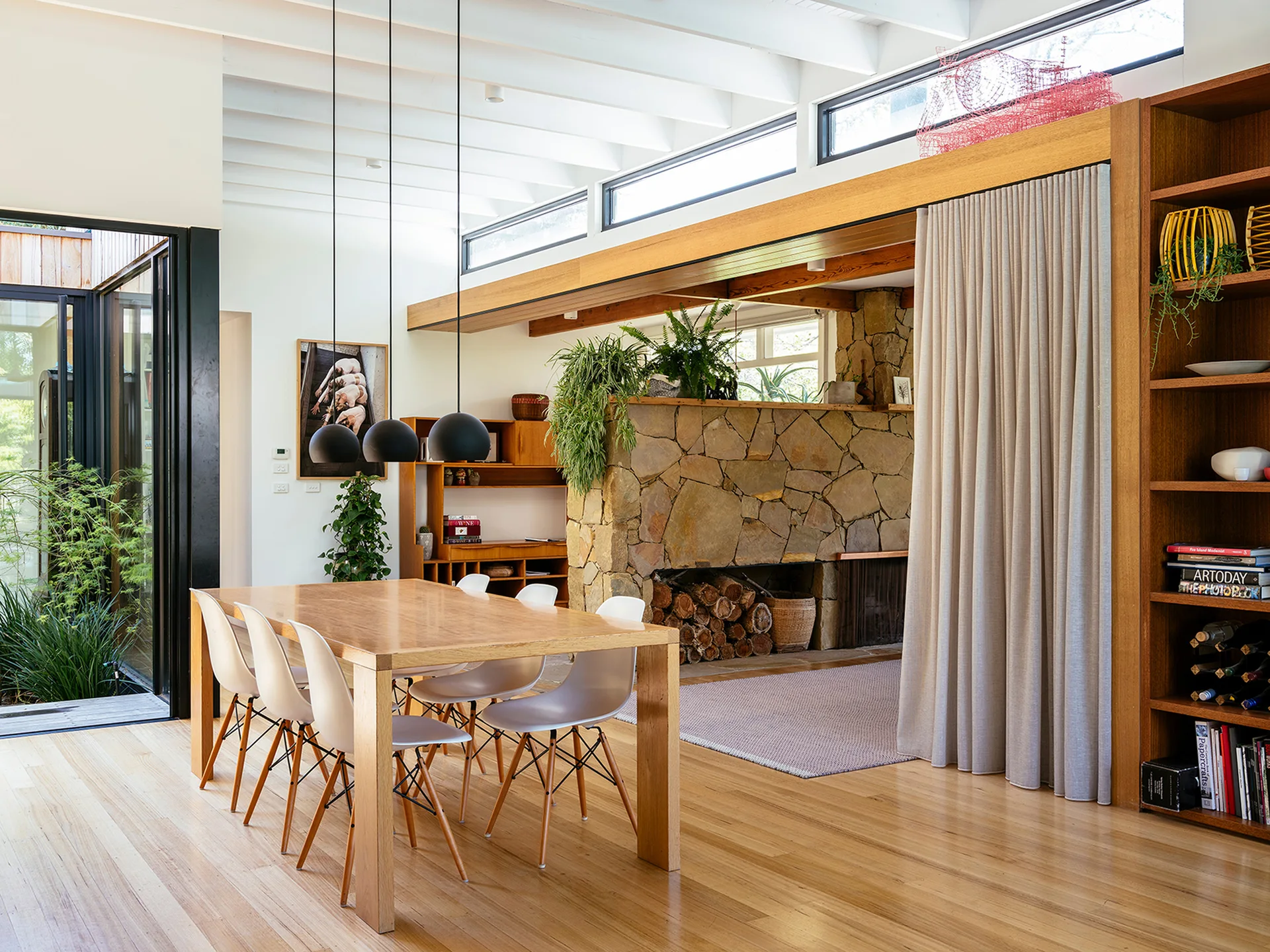 A bright dining room features a light wood table with white chairs, three black pendant lights, a floor-to-ceiling stone fireplace, and high ceilings.