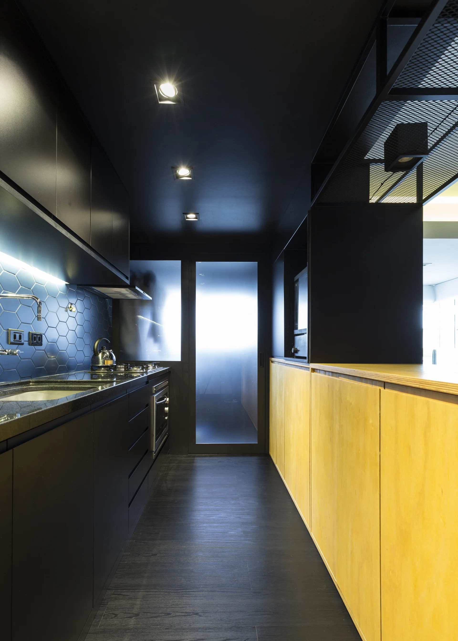 A galley kitchen features matte black cabinetry and a black hexagonal tile backsplash on one side, paired with warm wood-toned cabinets on the other.