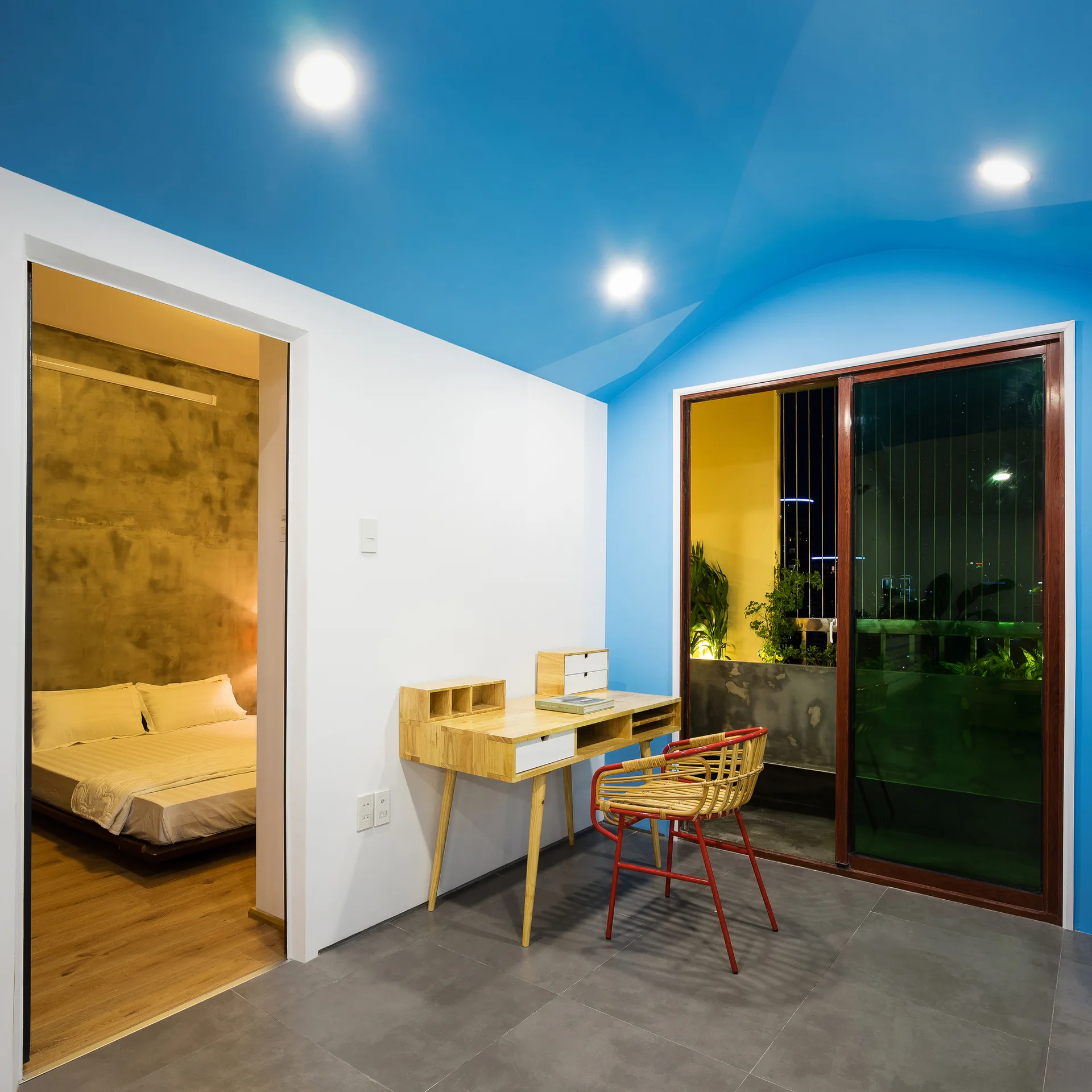 A home office with a wooden desk and rattan chair sits under a bright blue vaulted ceiling next to a bedroom.