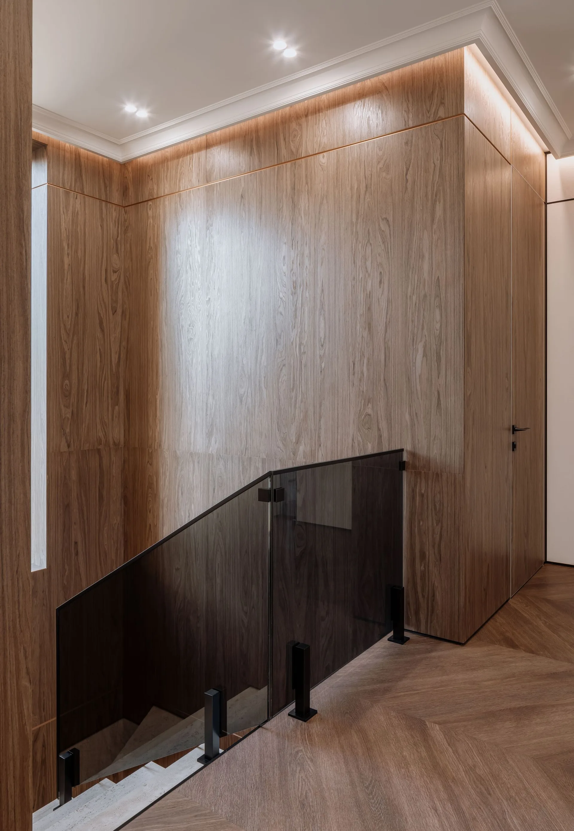 A modern staircase features warm wood-paneled walls, a dark tinted glass railing, chevron wood flooring, and glowing cove ceiling lighting.