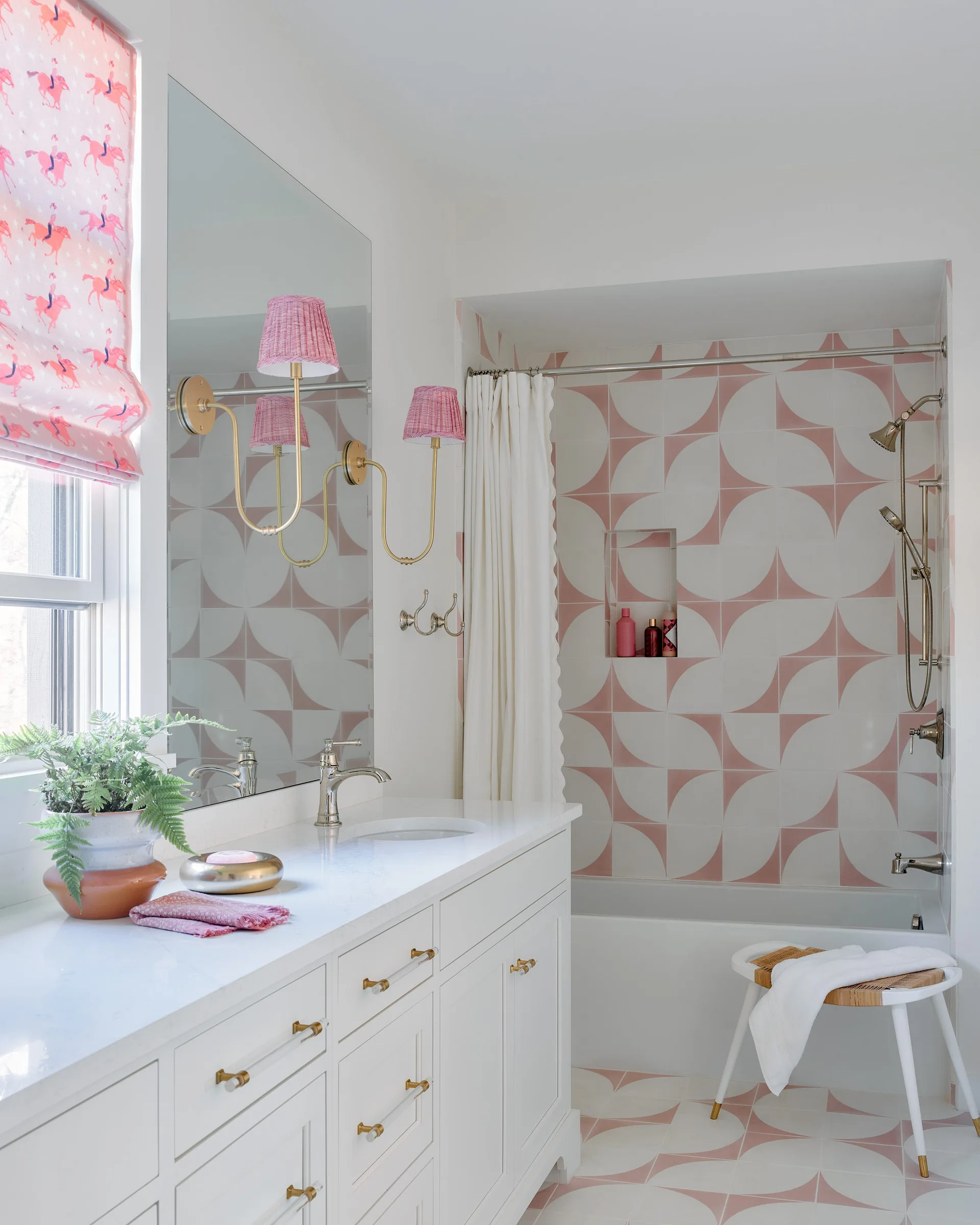 A bright bathroom features a white vanity, a large mirror with brass sconces, and a shower area with pink and white patterned geometric tiles.