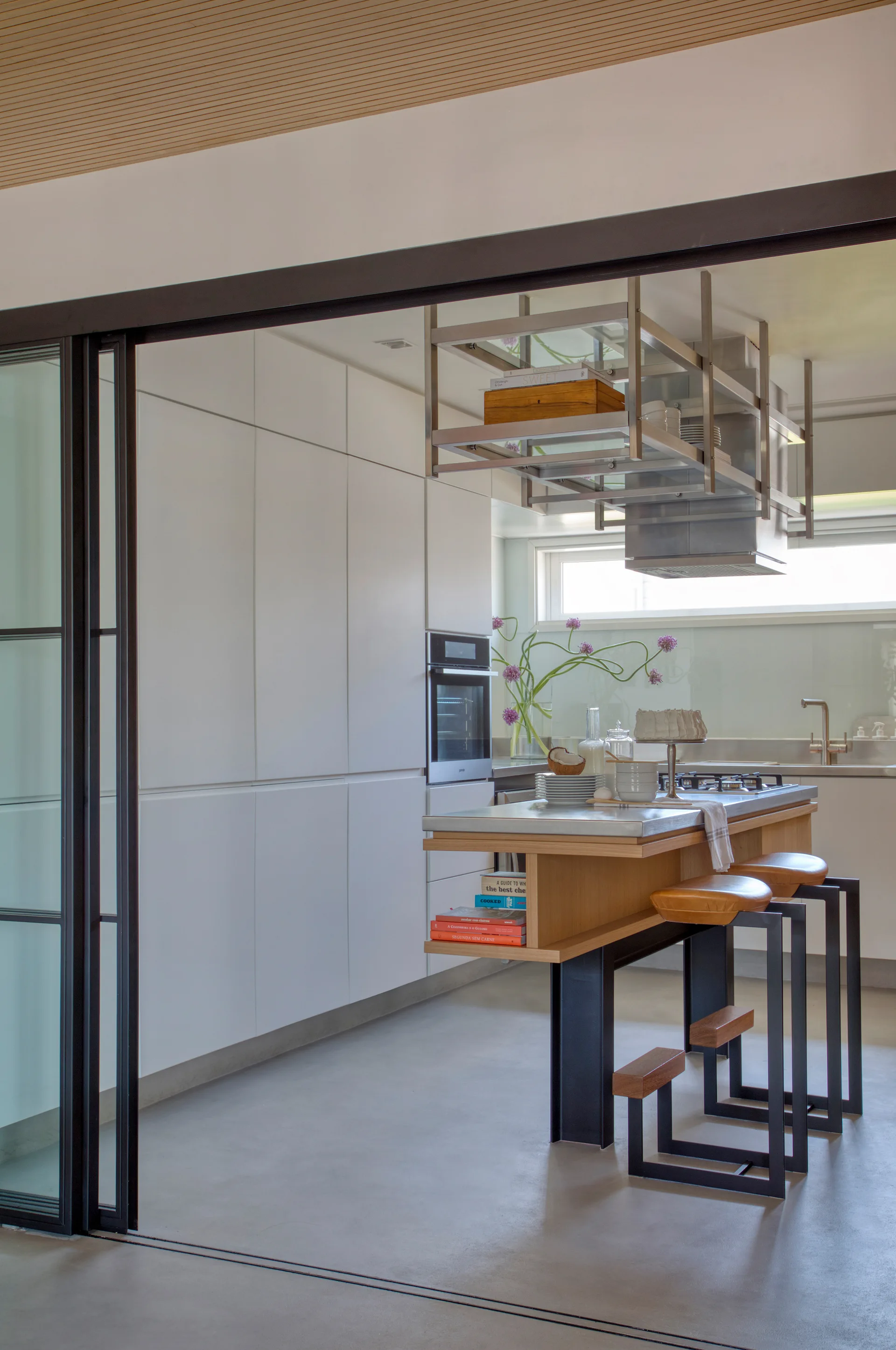 A modern kitchen with a wood slatted ceiling features white cabinetry, a stainless steel island with bar stools, and an overhead metal storage rack.