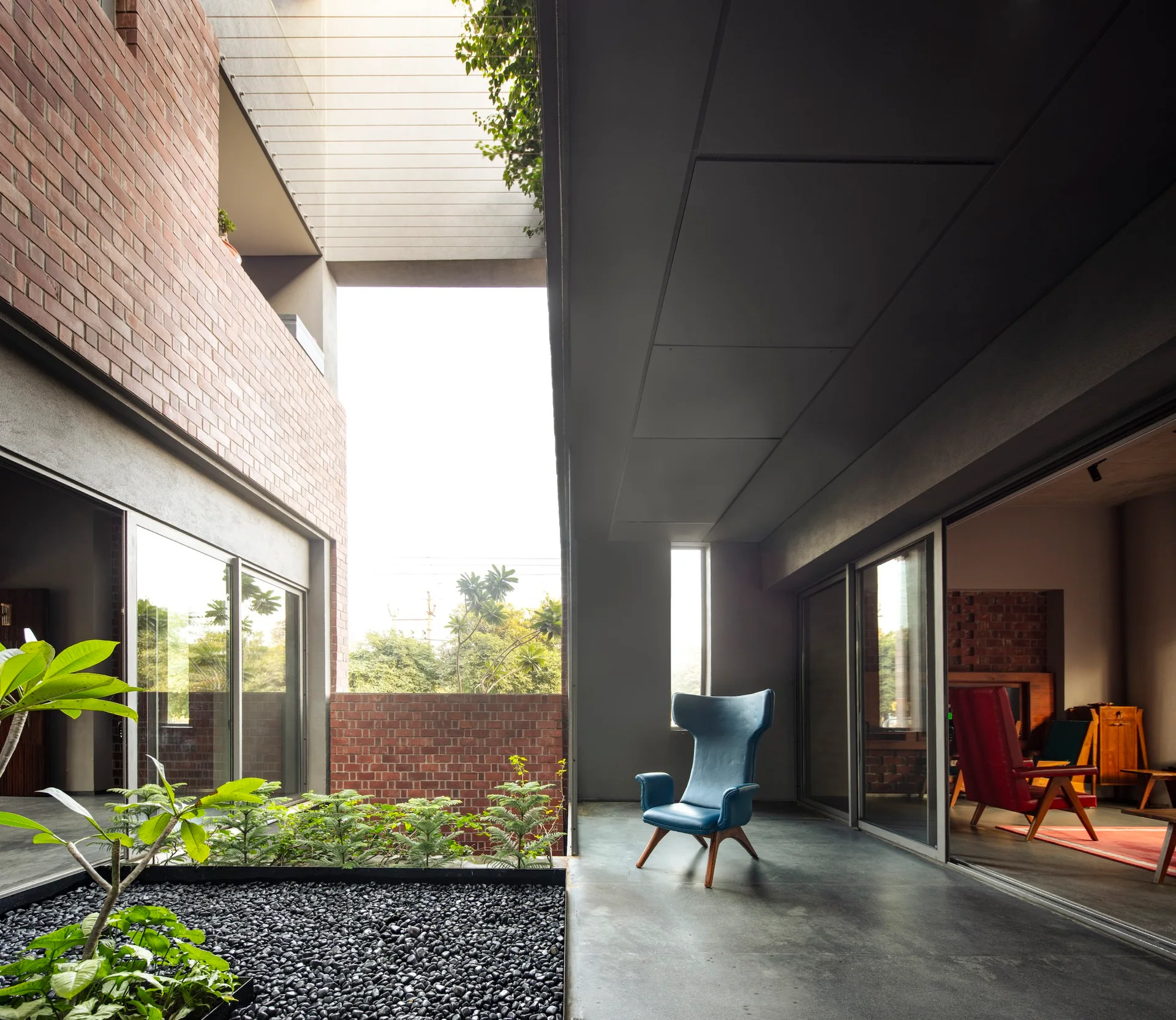 A modern covered concrete patio features a blue wingback armchair, sliding glass doors, and red brick walls next to a pebbled planter.