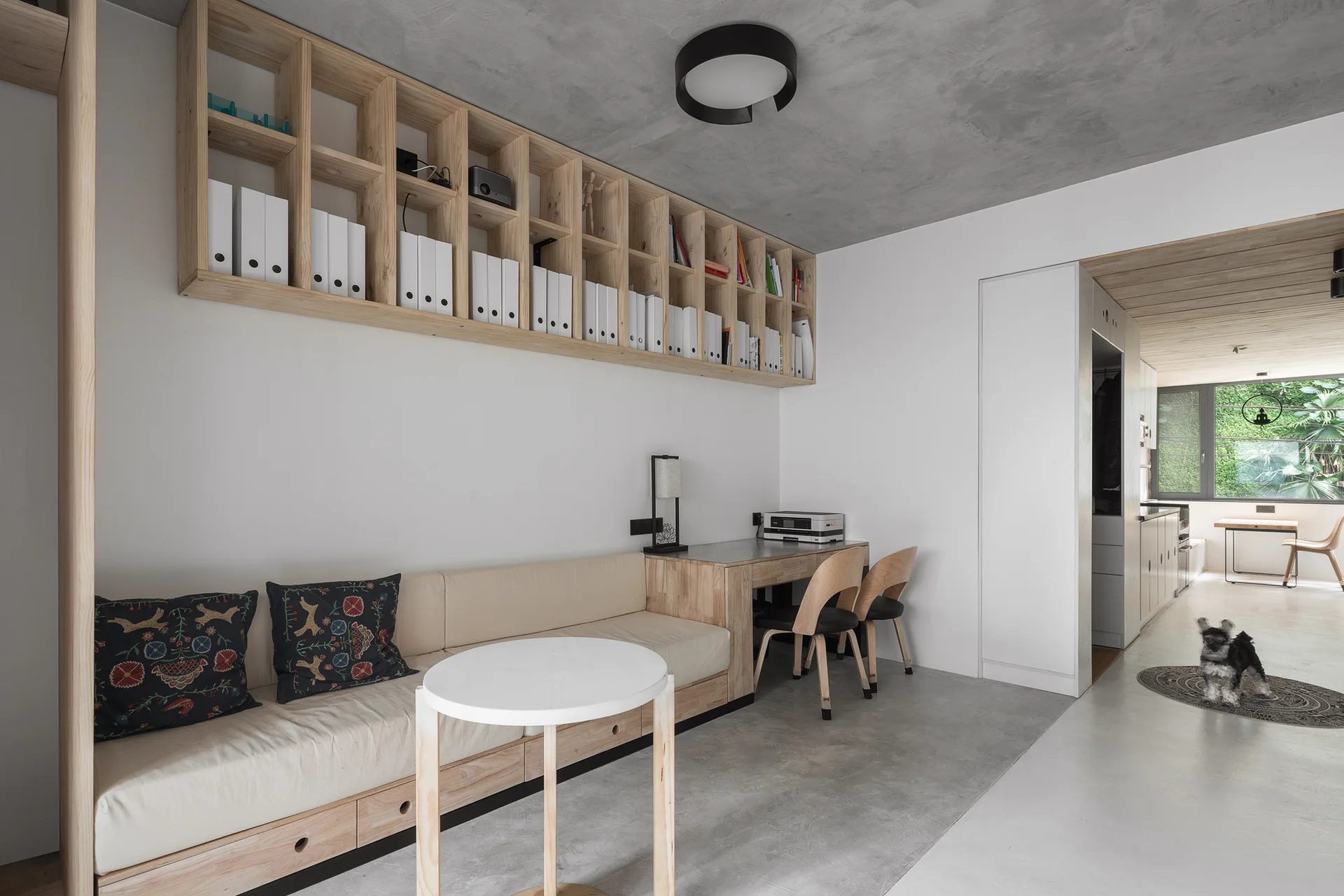 A modern home office featuring a built-in wooden desk and bench sofa under wall-mounted cubby shelving filled with white binders.