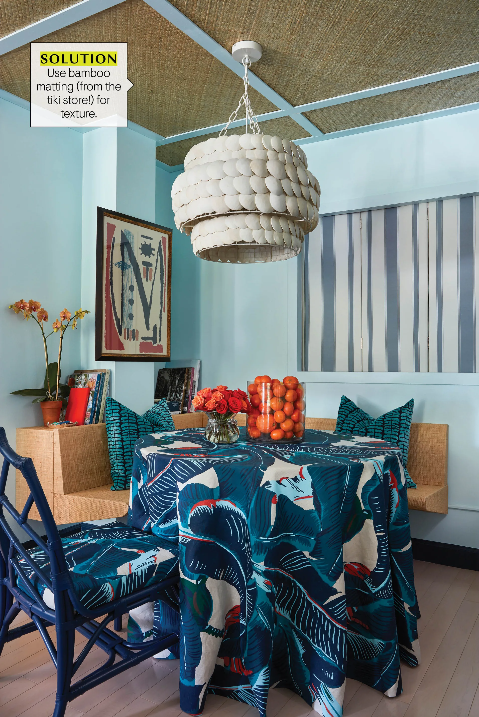 A dining nook features light blue walls, a woven corner banquette, a tropical leaf tablecloth, and a grid ceiling with bamboo matting.