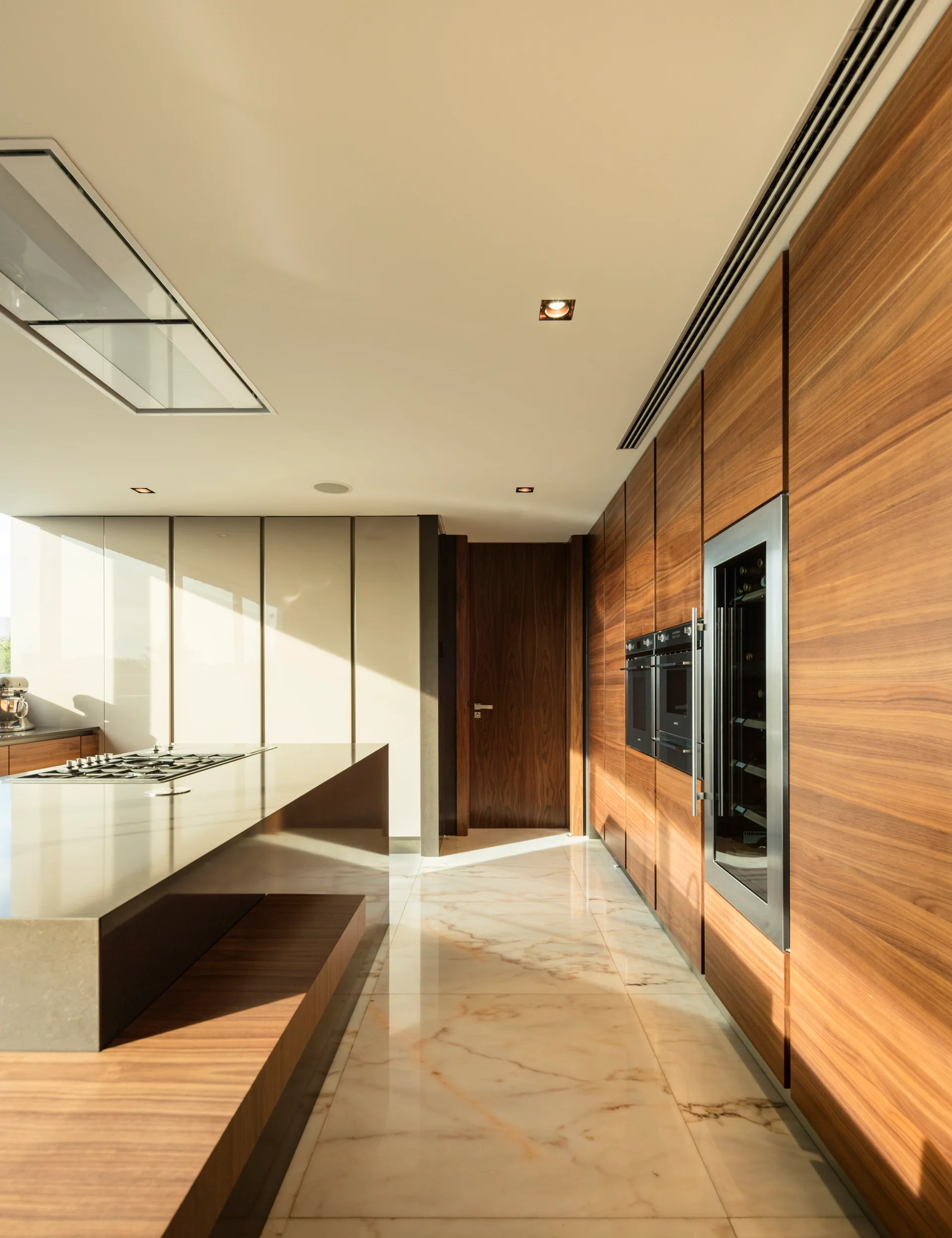 A modern kitchen featuring a sleek island with a cooktop, a wall of wood veneer cabinetry, integrated steel appliances, and polished marble floors.