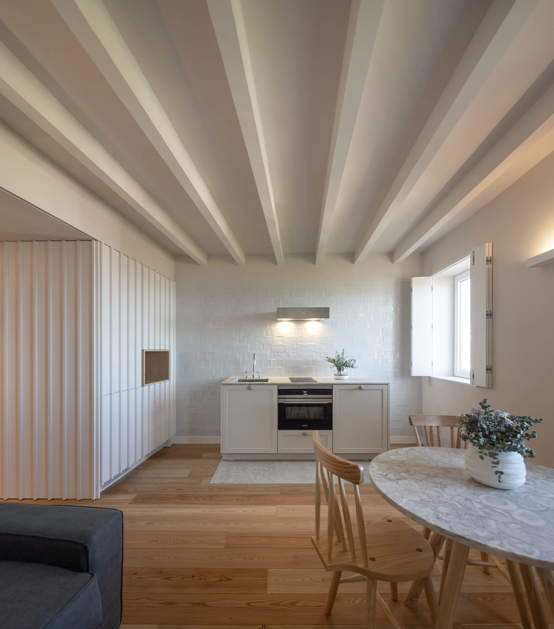 A bright kitchen and dining area features white ceiling beams, light wood floors, a round stone table, and minimal off-white cabinets.