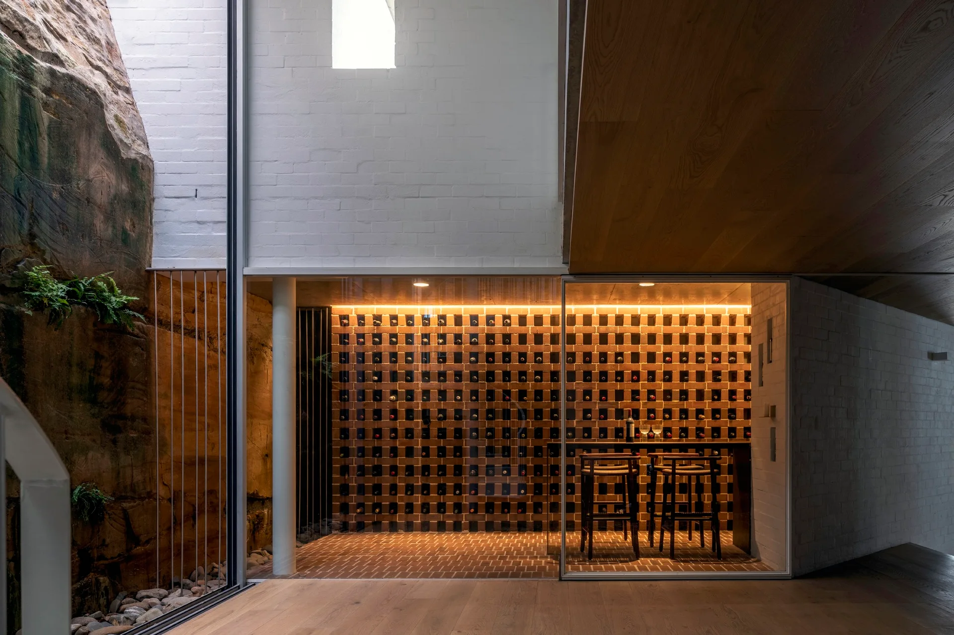 A glass-enclosed modern wine cellar with a terracotta brick bottle grid, wooden counter, bar stools, and an adjacent natural rock face lightwell.