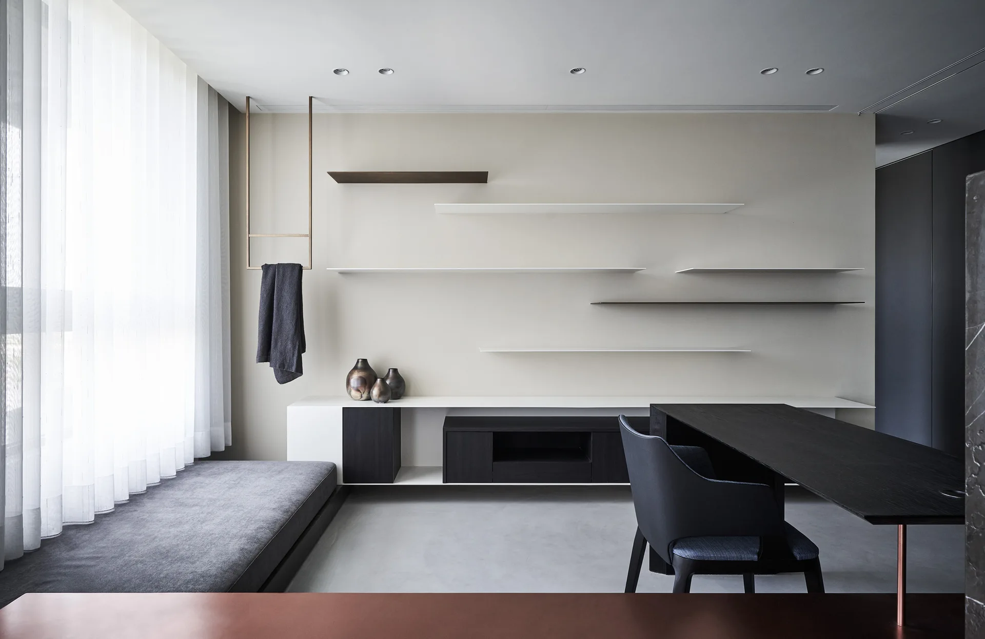 A minimalist living area features staggered floating shelves on a beige wall, a low built-in console, and an upholstered gray bench next to a window.