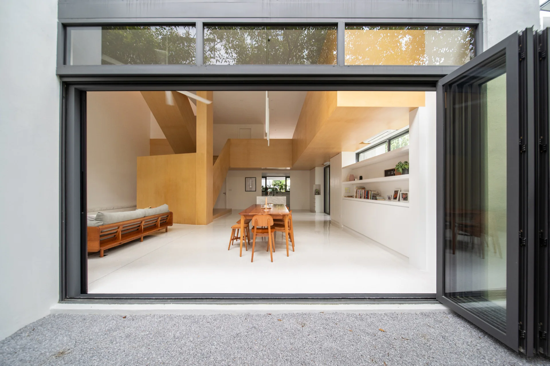 An open plan living area features white floors, a wooden dining table, a massive natural wood staircase structure, and open bifold doors.