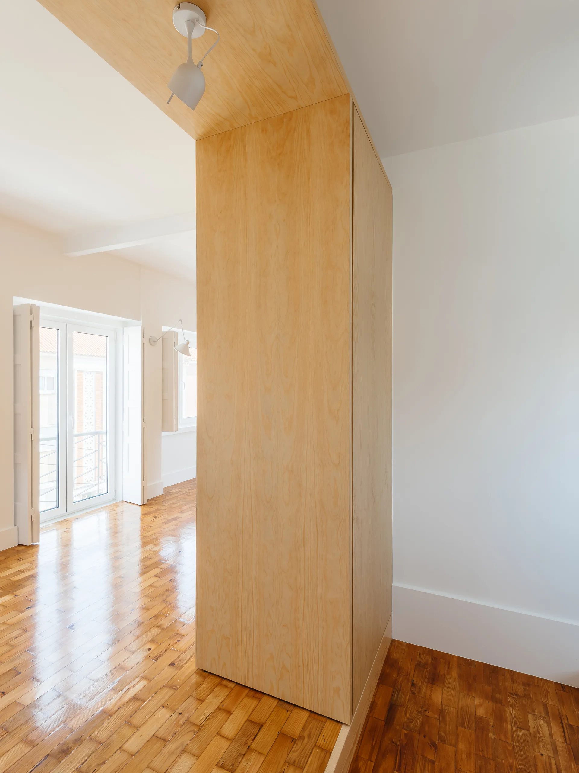 A minimalist room featuring a large light wood partition cabinet, polished parquet flooring, and white shuttered windows.