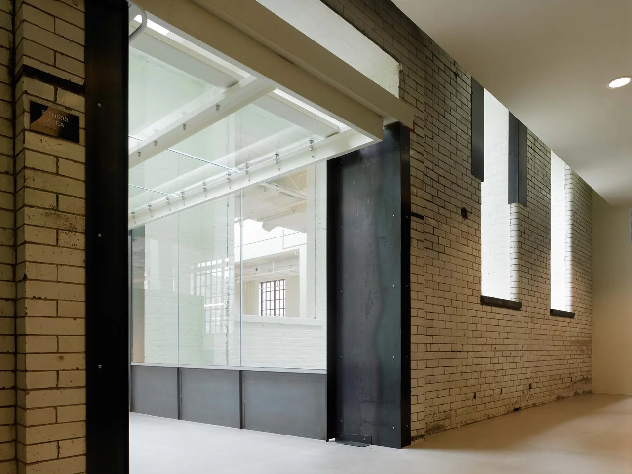 A hallway with painted white brick walls and recessed windows leads past a large glass and black steel partition.
