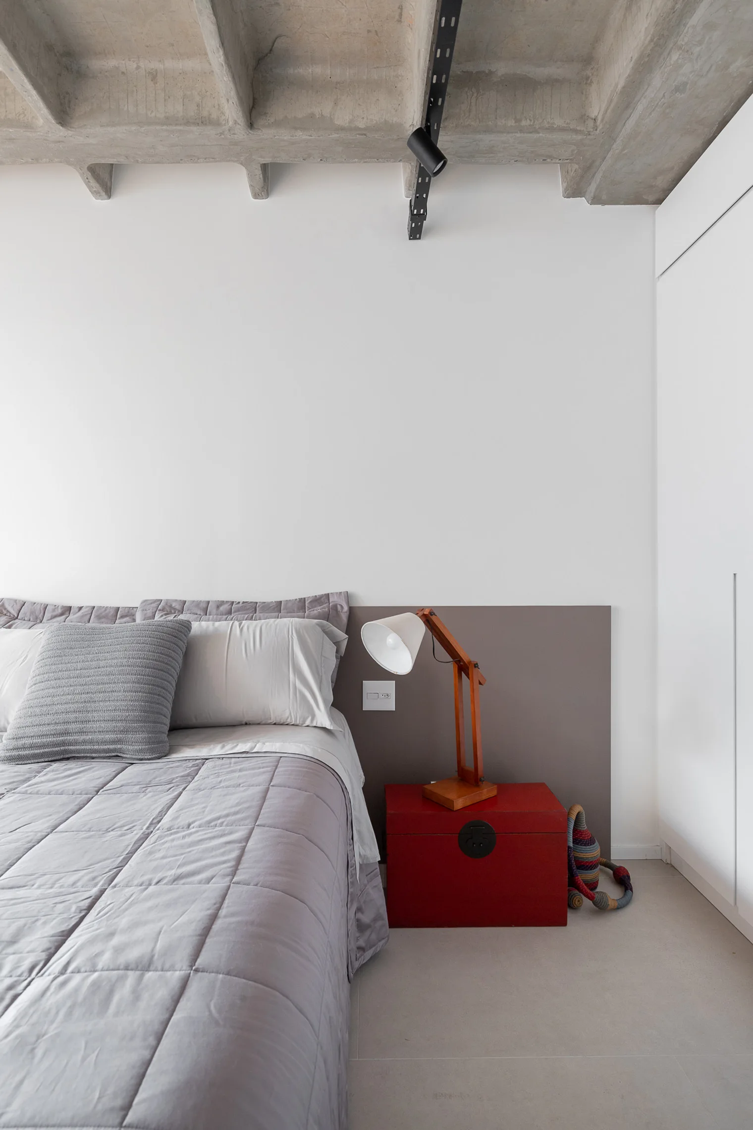 A modern bedroom features a gray quilted bed, an articulated wood desk lamp on a red chest nightstand, and an exposed concrete waffle ceiling.