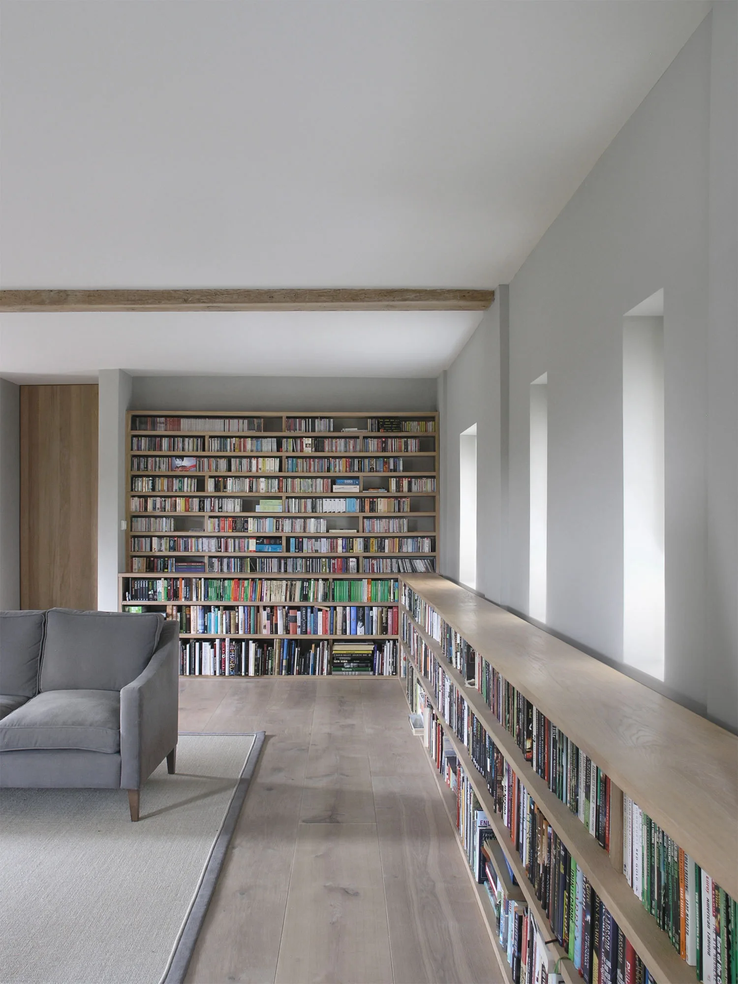 A minimalist grey room features a large built-in wooden bookcase, a low shelf under deep recessed windows, and a grey sofa on a matching rug.