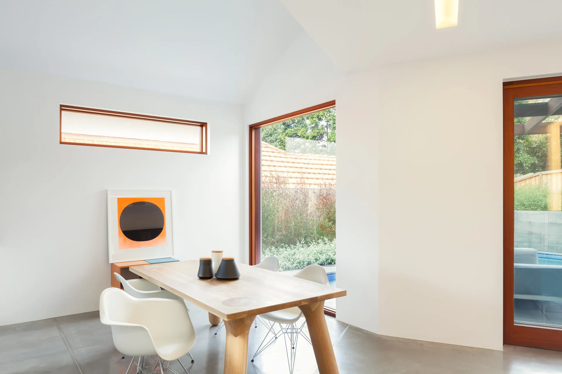 A modern minimalist dining room featuring a light wood table, white shell chairs, concrete floors, and wood-framed glass doors leading to a garden.