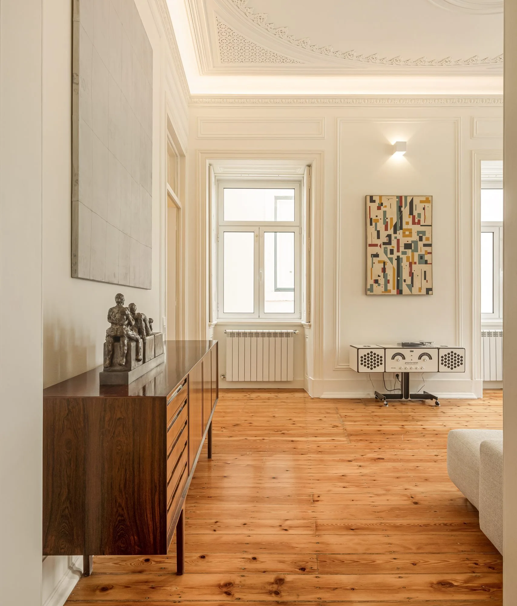 A spacious room with warm pine floors features a mid-century wood sideboard, a vintage record console, and ornate neoclassical wall and ceiling moldings.