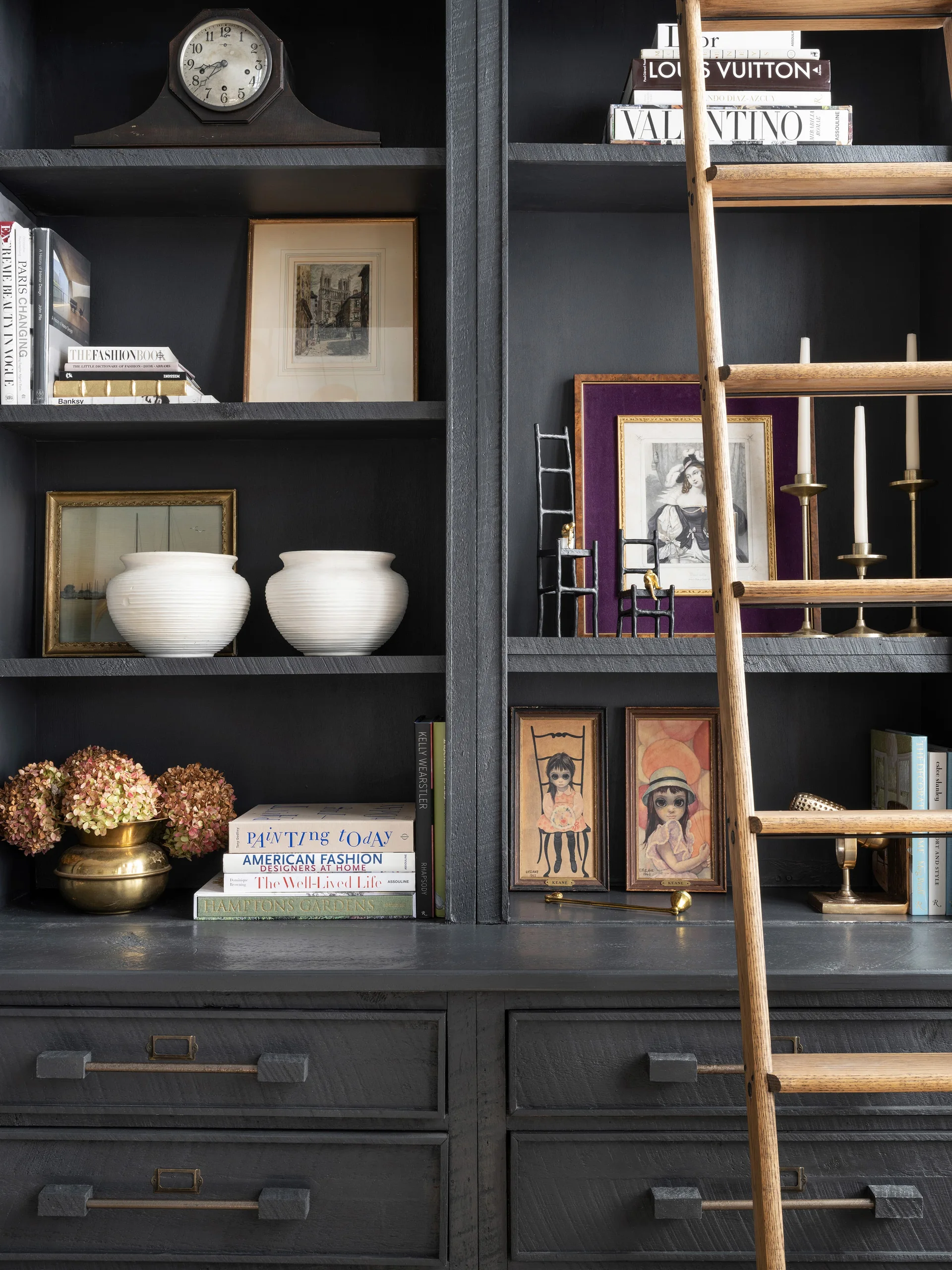 A dark charcoal painted built-in bookcase styled with books, art, and ceramic vases features a wooden rolling library ladder.