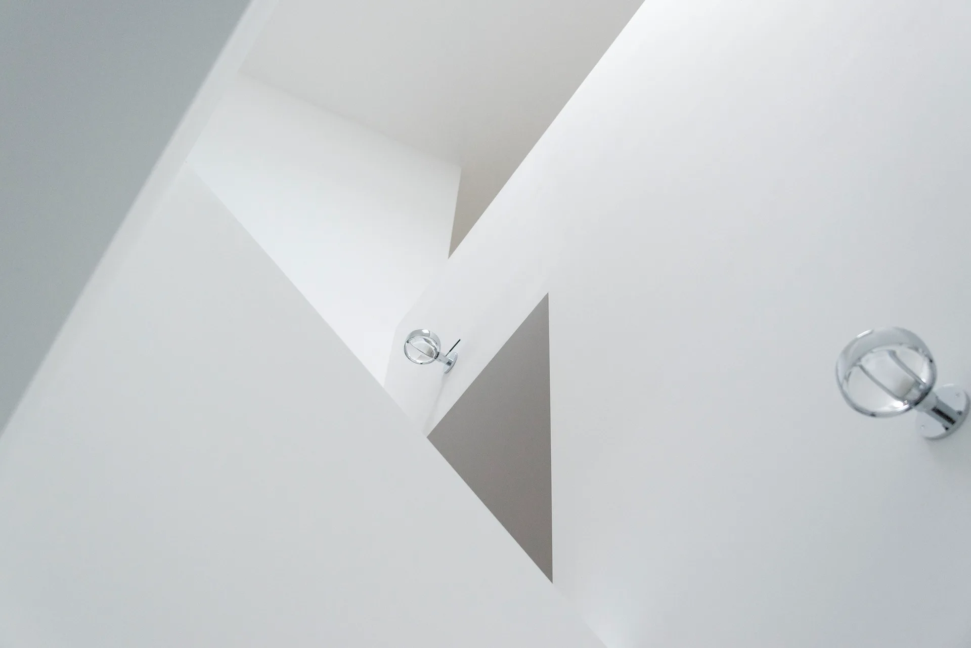 An architectural view of a minimalist white stairwell featuring geometric intersecting wall planes and two round chrome sconces.