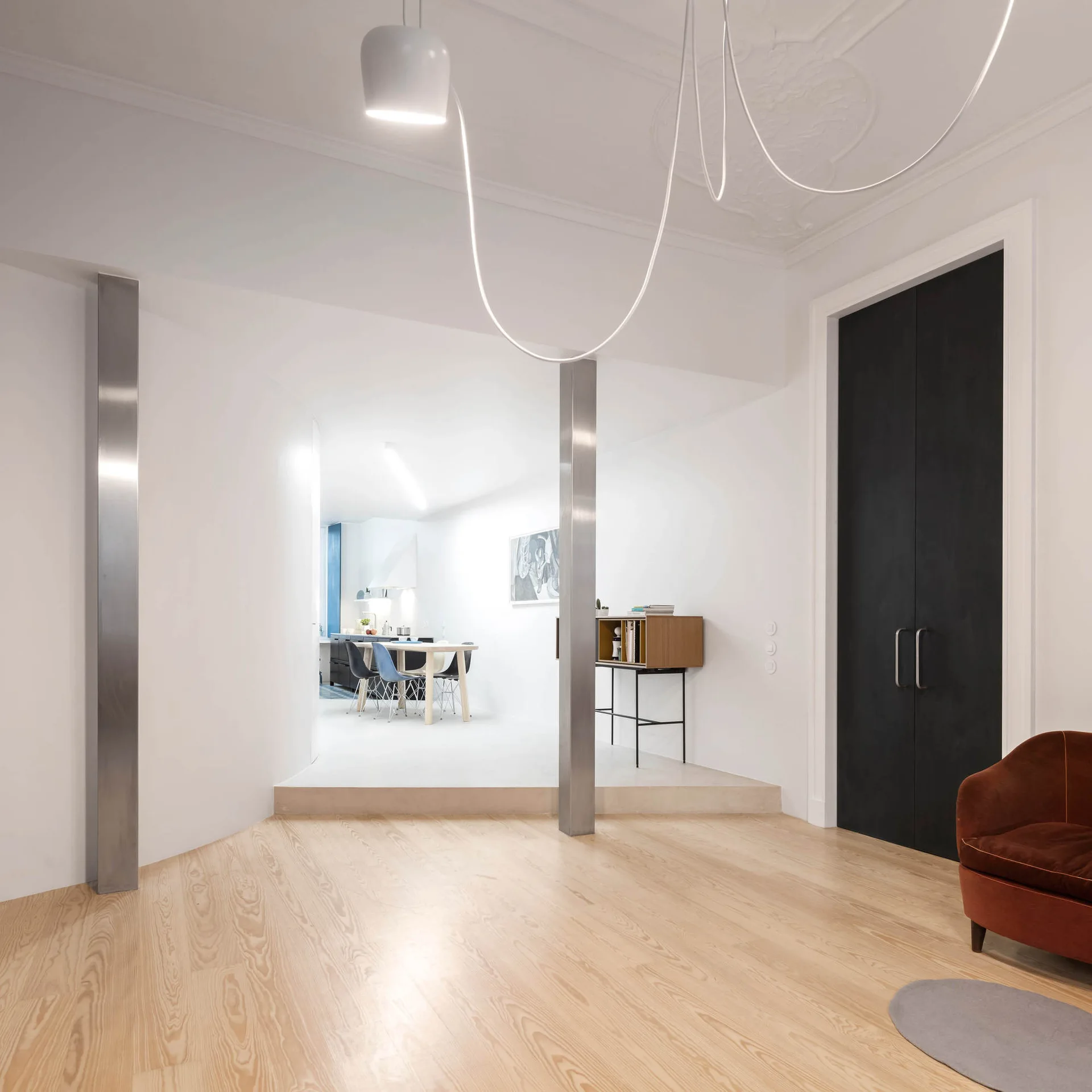 A modern split-level living space with light wood flooring, brushed metal columns, tall black double doors, and a draped white pendant light.