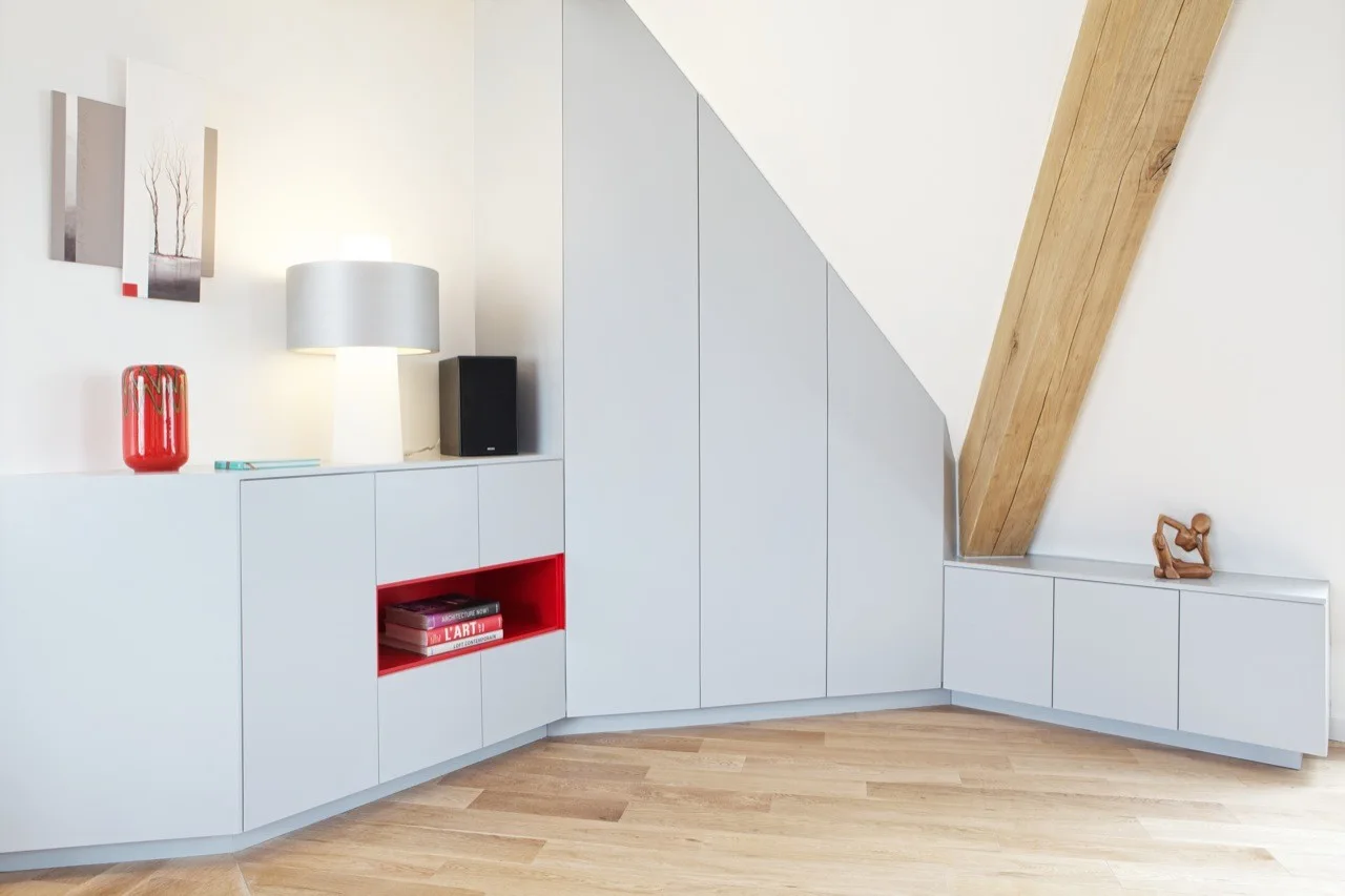 Custom light grey cabinets fitted under a sloped attic ceiling next to an exposed wooden beam and light wood flooring.