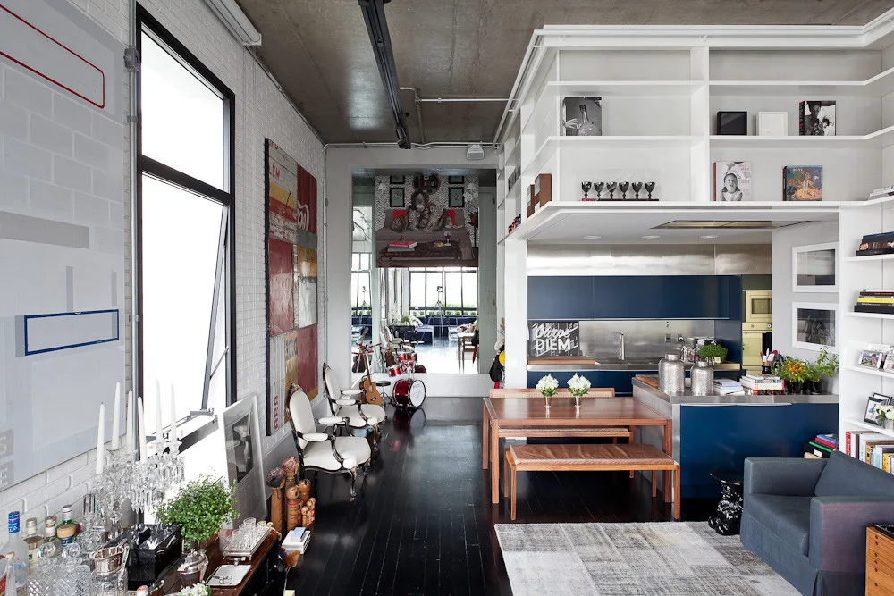 An open-plan industrial loft featuring a wooden dining table, white painted brick walls, a blue kitchen, and built-in shelving.