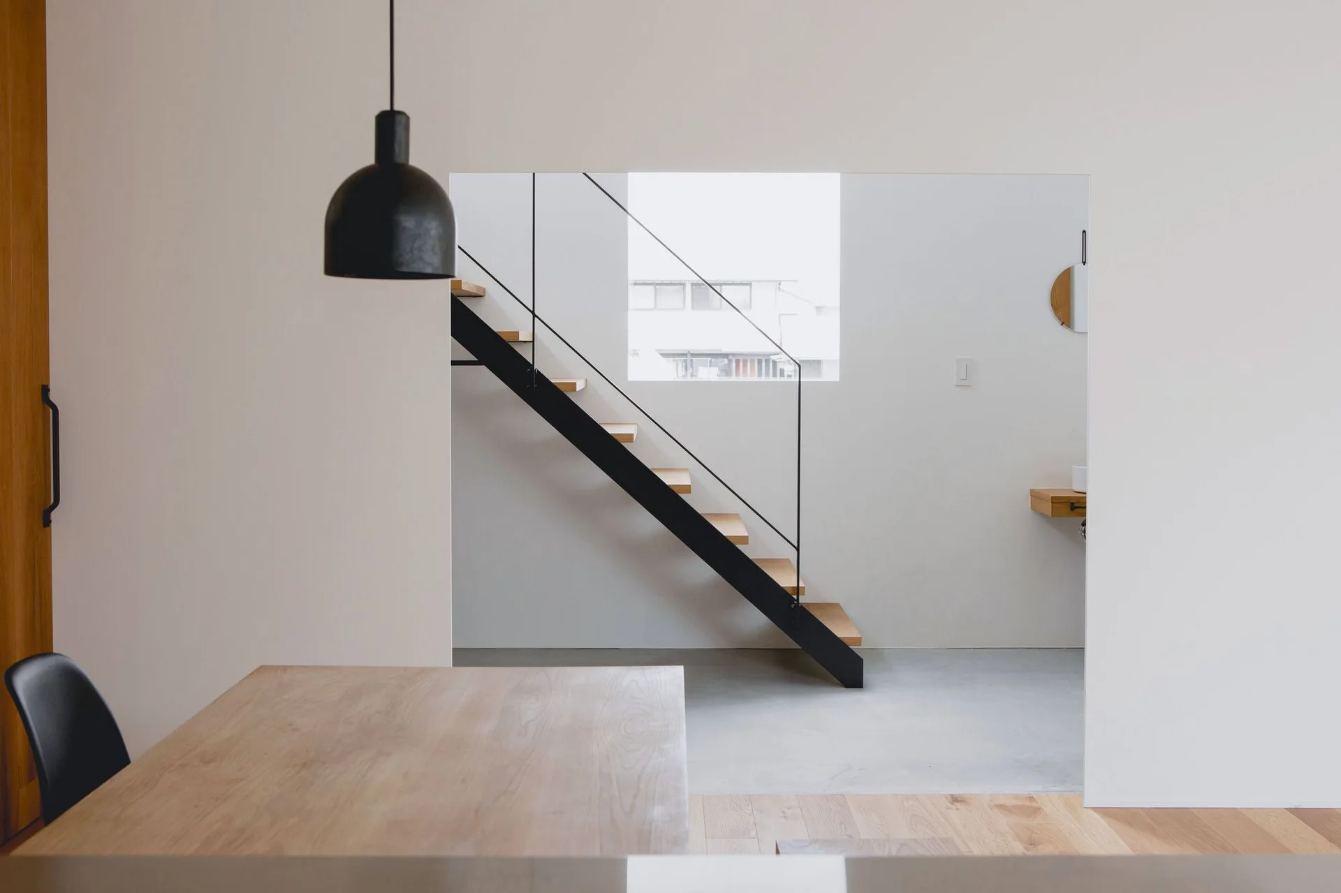 A view through a wall opening toward a minimalist black metal floating staircase with wood treads next to a bright window.