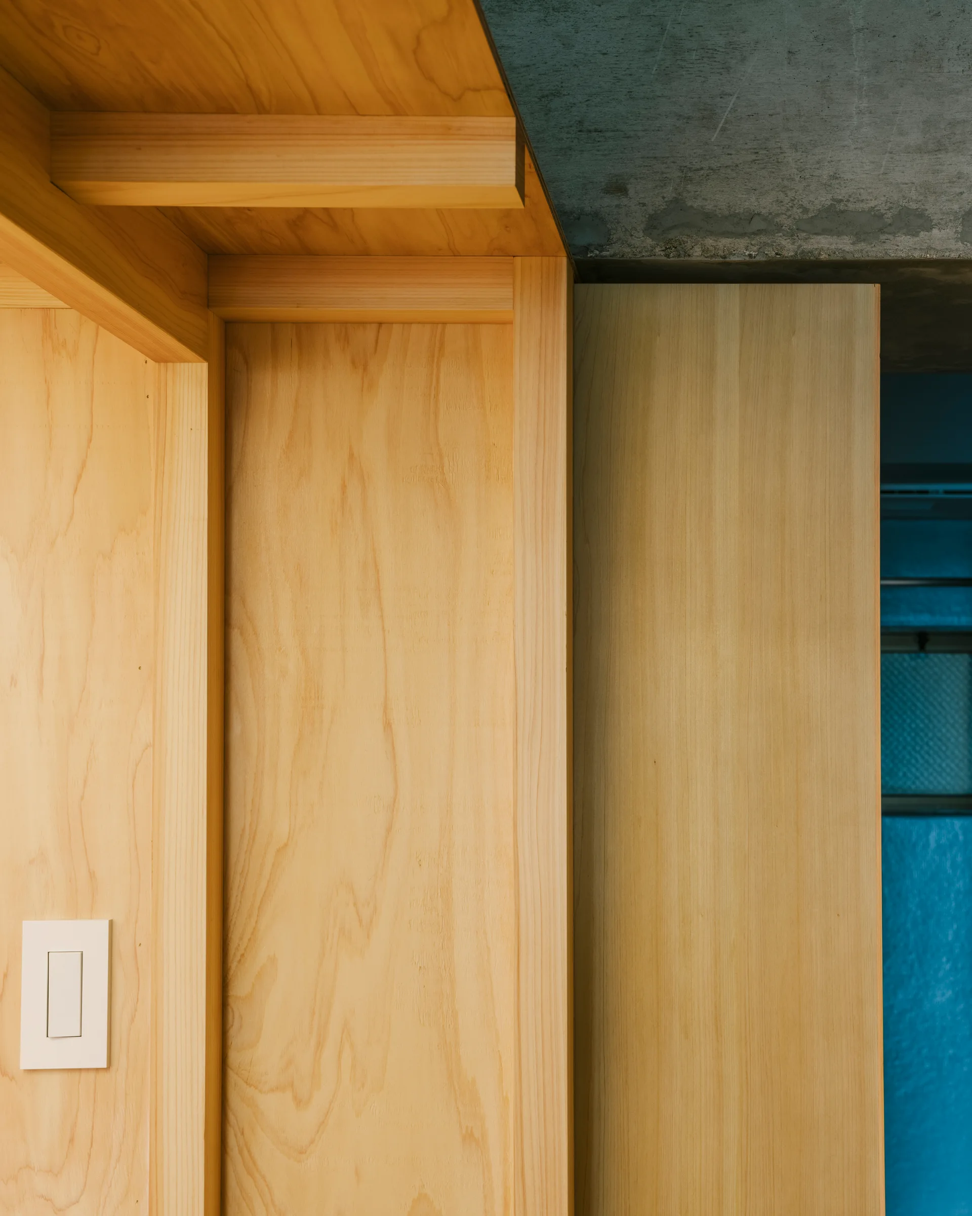 A close-up of light-toned plywood wall paneling and custom joinery meeting a textured gray concrete ceiling, featuring a simple white light switch.