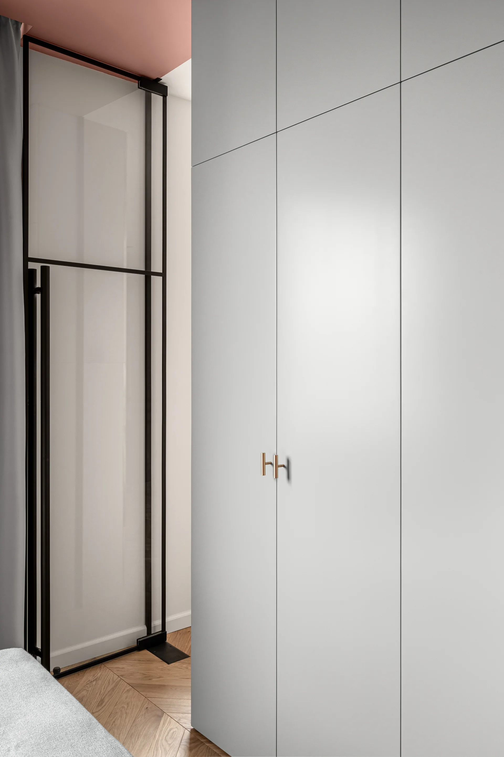 A pale grey built-in closet with copper handles stands next to a black-framed glass partition on herringbone wood flooring with a pink ceiling accent.