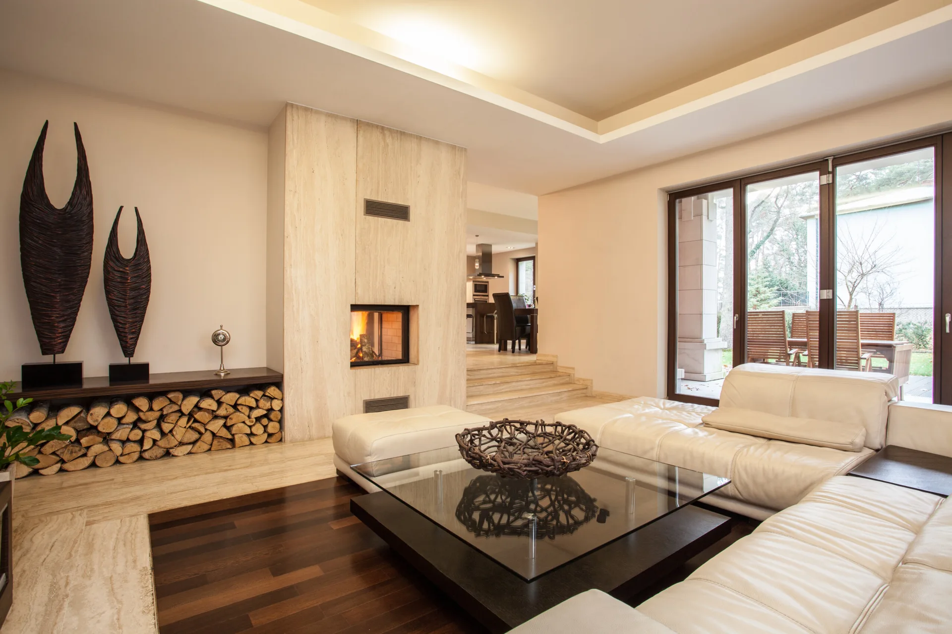 A modern living room with a white leather sectional sofa, a stone-clad fireplace, dark wood flooring, and a low bench storing stacked logs.